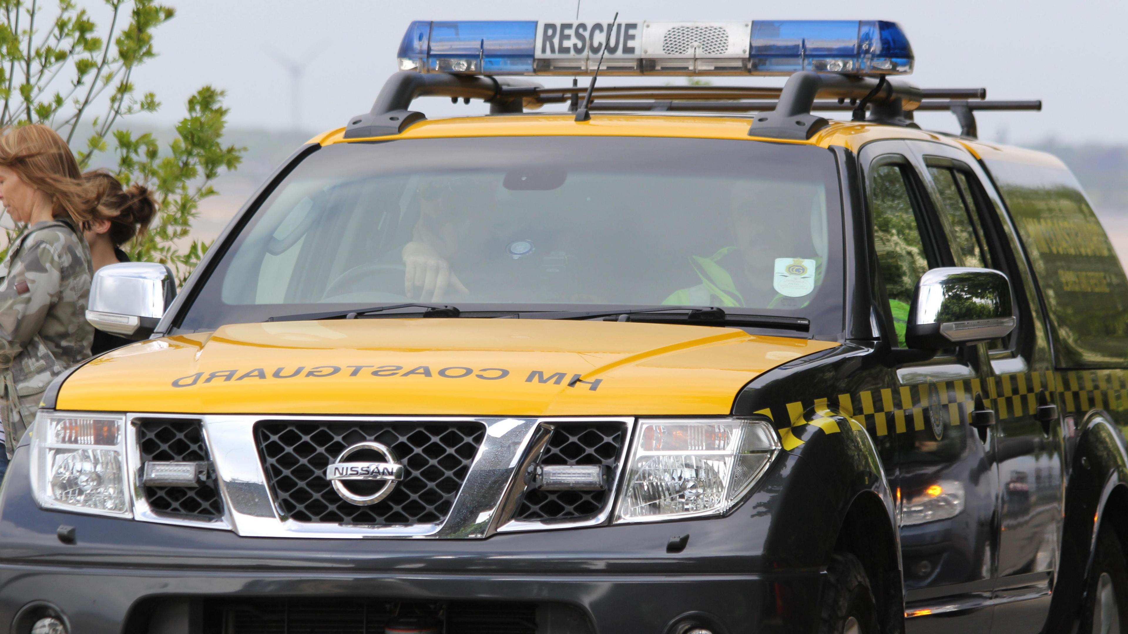 A yellow and black HM Coastguard pick-up truck at the scene of an incident. Two people are sitting inside the vehicle. Two women are stood next to the vehicle. One of the woman has a camouflage jacket on.