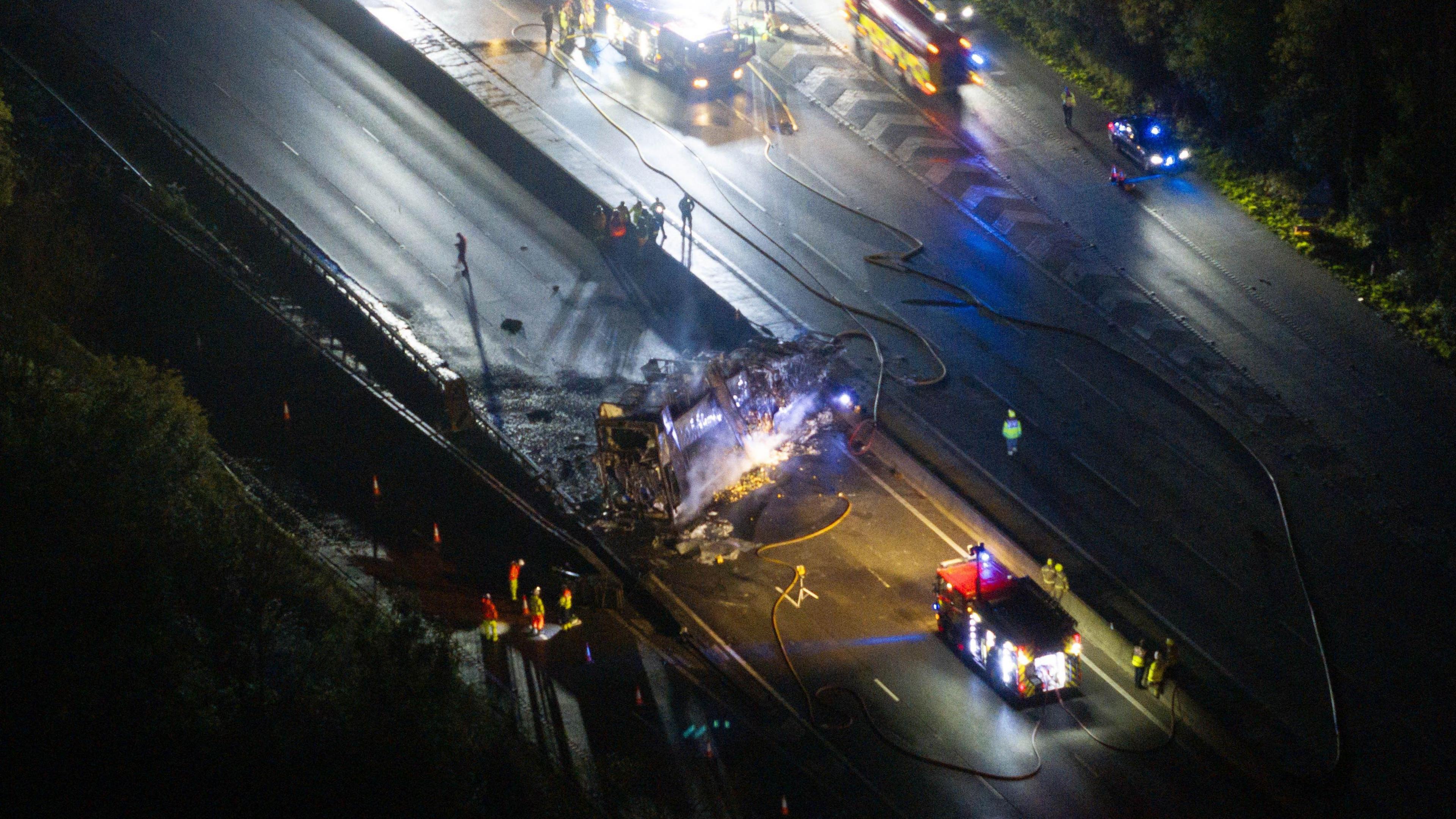 An aerial shot of a burnt out vehicle on a section of motorway. Fire hoses are visible.