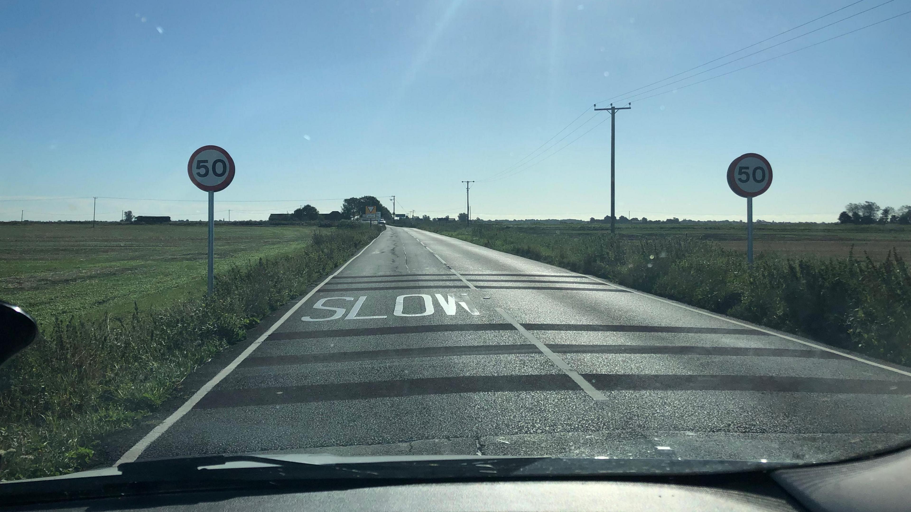 Two 50mph speed limit signs on the sides of the B1093 road, with SLOW painted on the road surface as vehicles approach the Boots Bridge in the distance