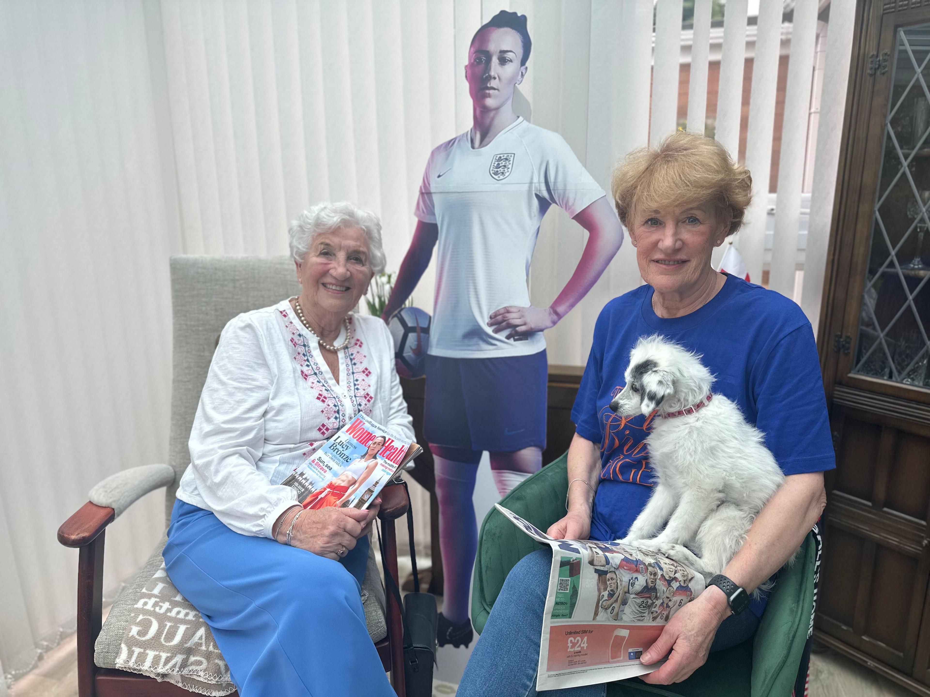 Gloria Stapleton (left) has short white hair and is smiling. She wears a pearl necklace and white cardigan. She is holding a copy of Women's Health magazine, which has a picture of Lucy Bronze on it. Julie Tough (right) has short ginger hair and holds a small white dog and a newspaper. A cardboard cutout of Lucy is behind them.
