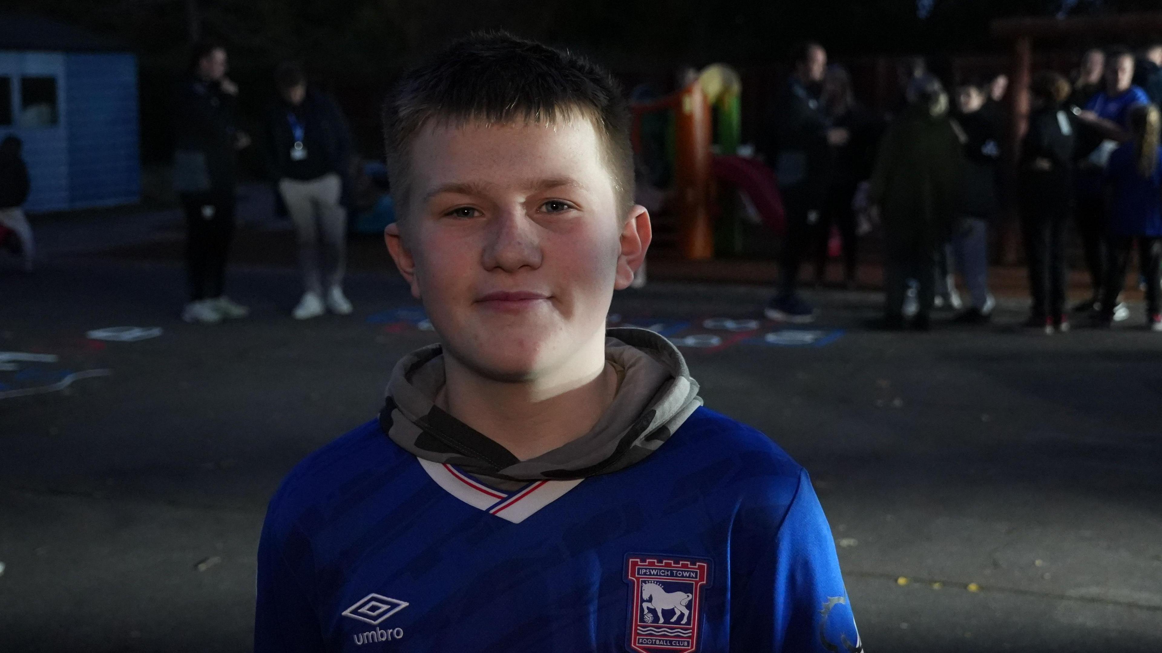 Archie smiles at the camera while standing outside a hub at night with several other people. He has short dark hair and wears a grey hoodie and Ipswich Town shirt