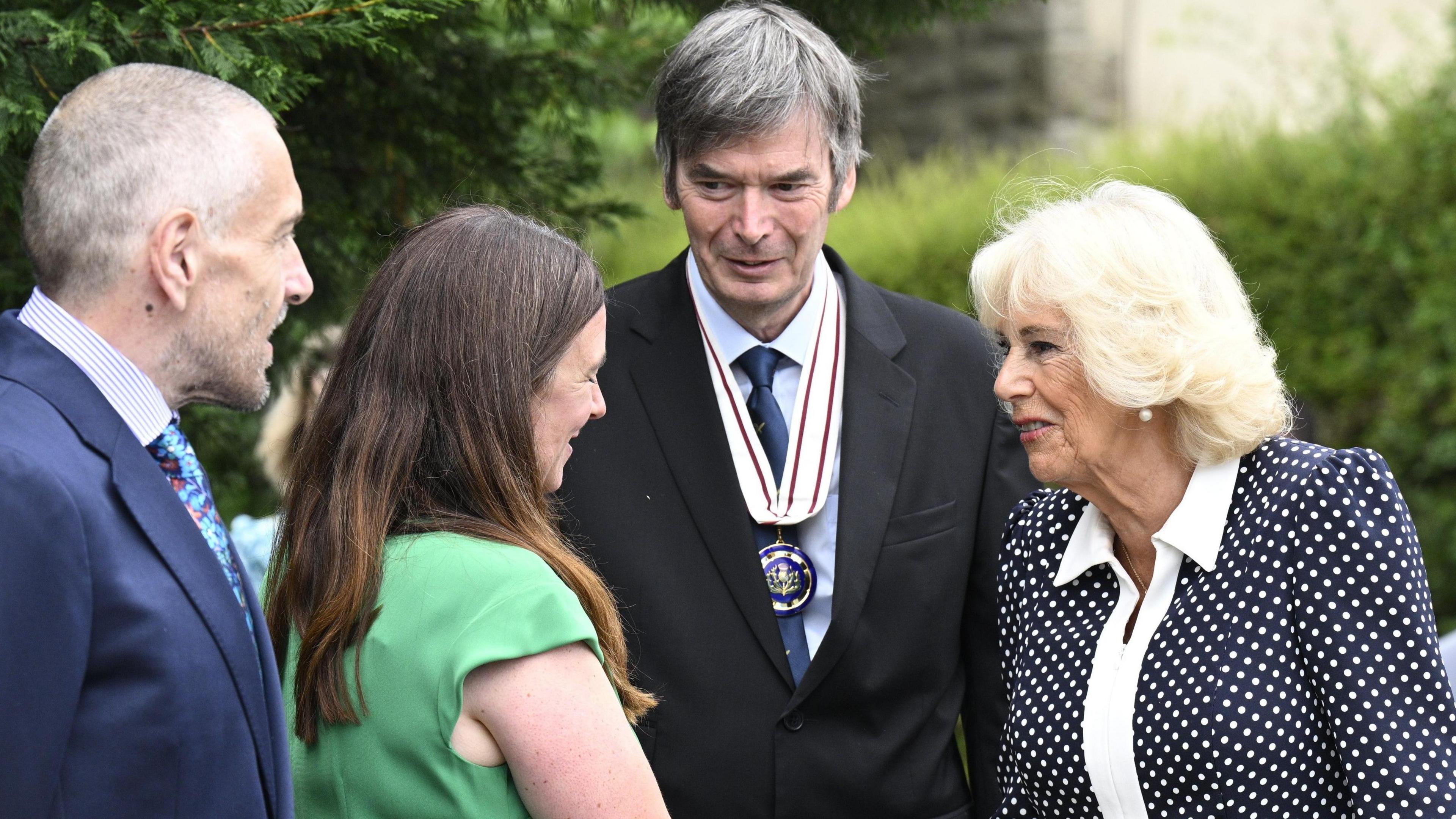 A man in a navy blue suit and colourful tie and a woman with long brown hair and a green dress greet writer Sir Ian Rankin, who wears a black suit with a navy blue tie and official medal, and Queen Camilla, wearing a black and white polka-dot dress with a white collar.