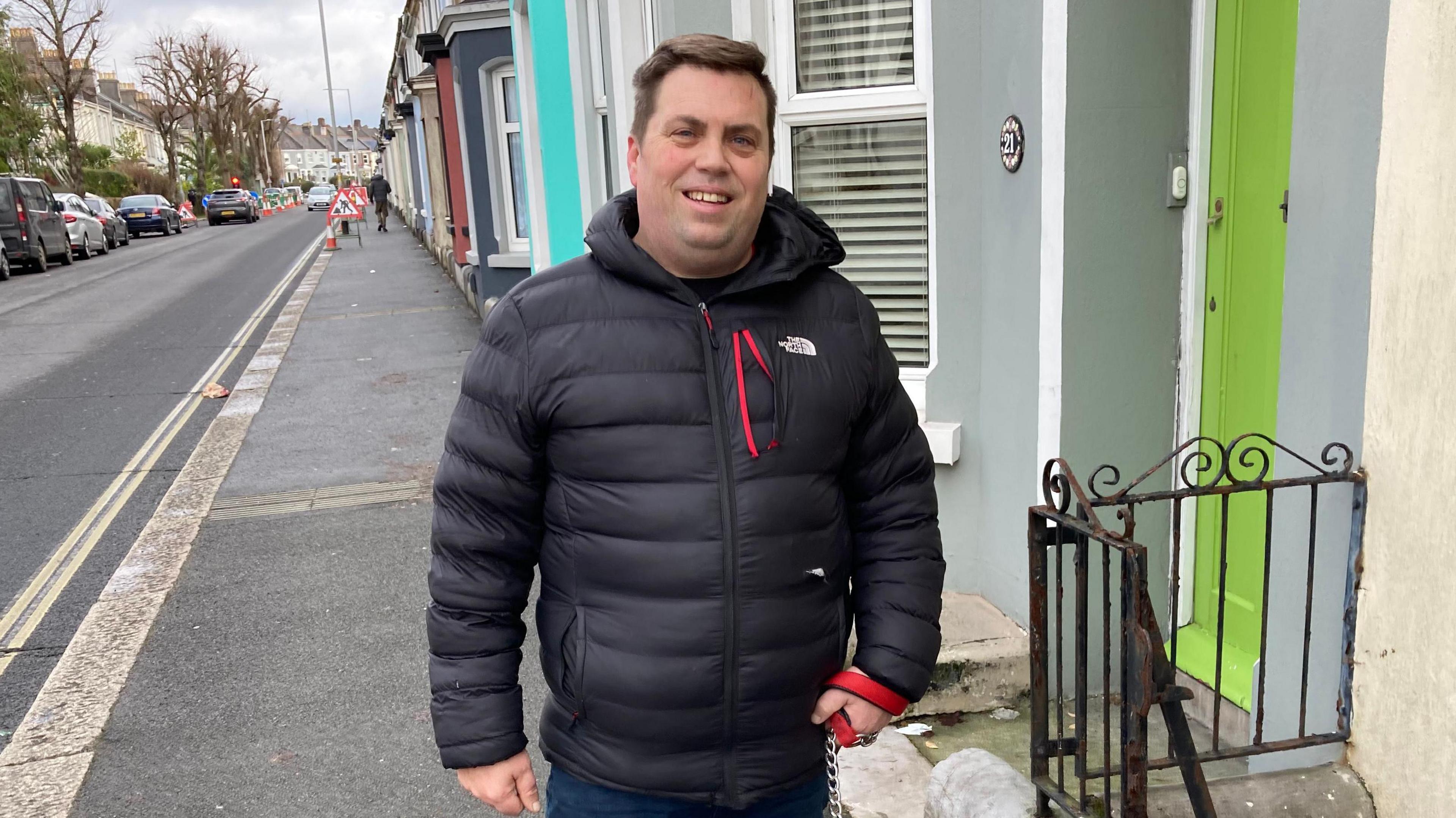 Roger Smith wearing a black North Face coat stands on a street outside a row of terrace houses. The door next to him is lime green.