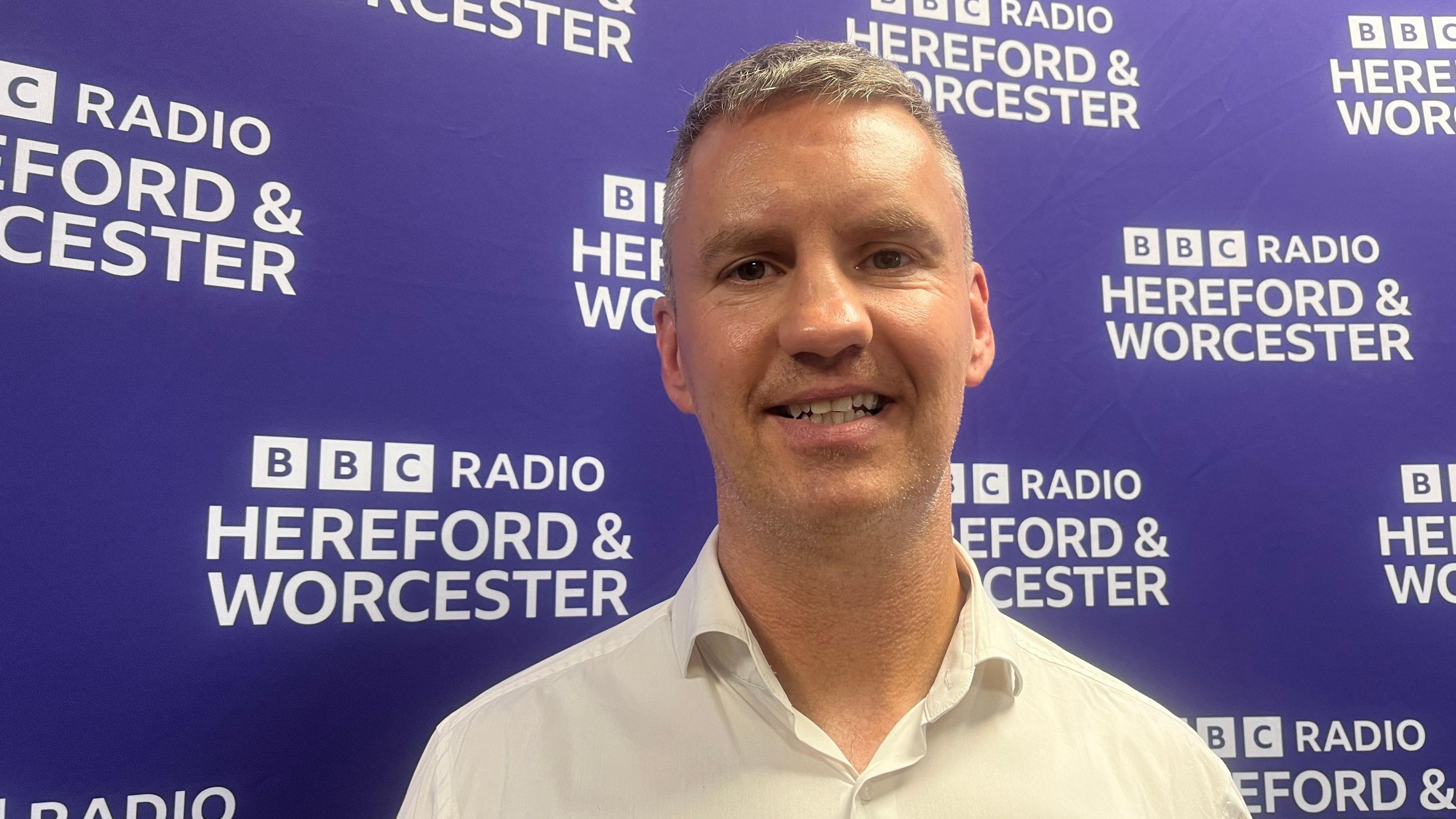 Stephen Vaughan stands in front of a purple board with BBC Hereford and Worcester displayed in white letters. He is smiling and wearing an open-necked white shirt