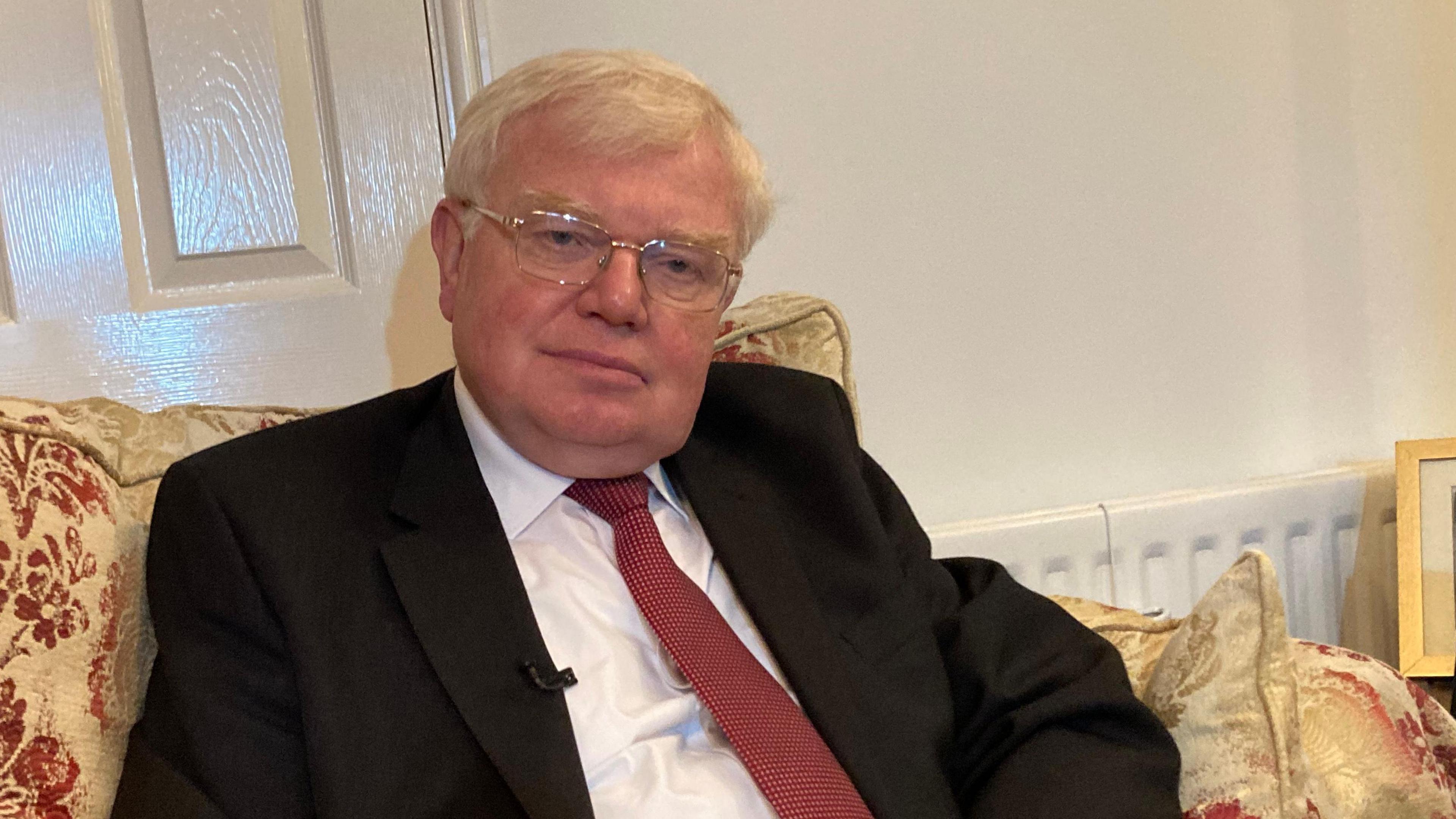 Dr Tim Brain, wearing a dark suit and red tie, sitting at home on a sofa. 