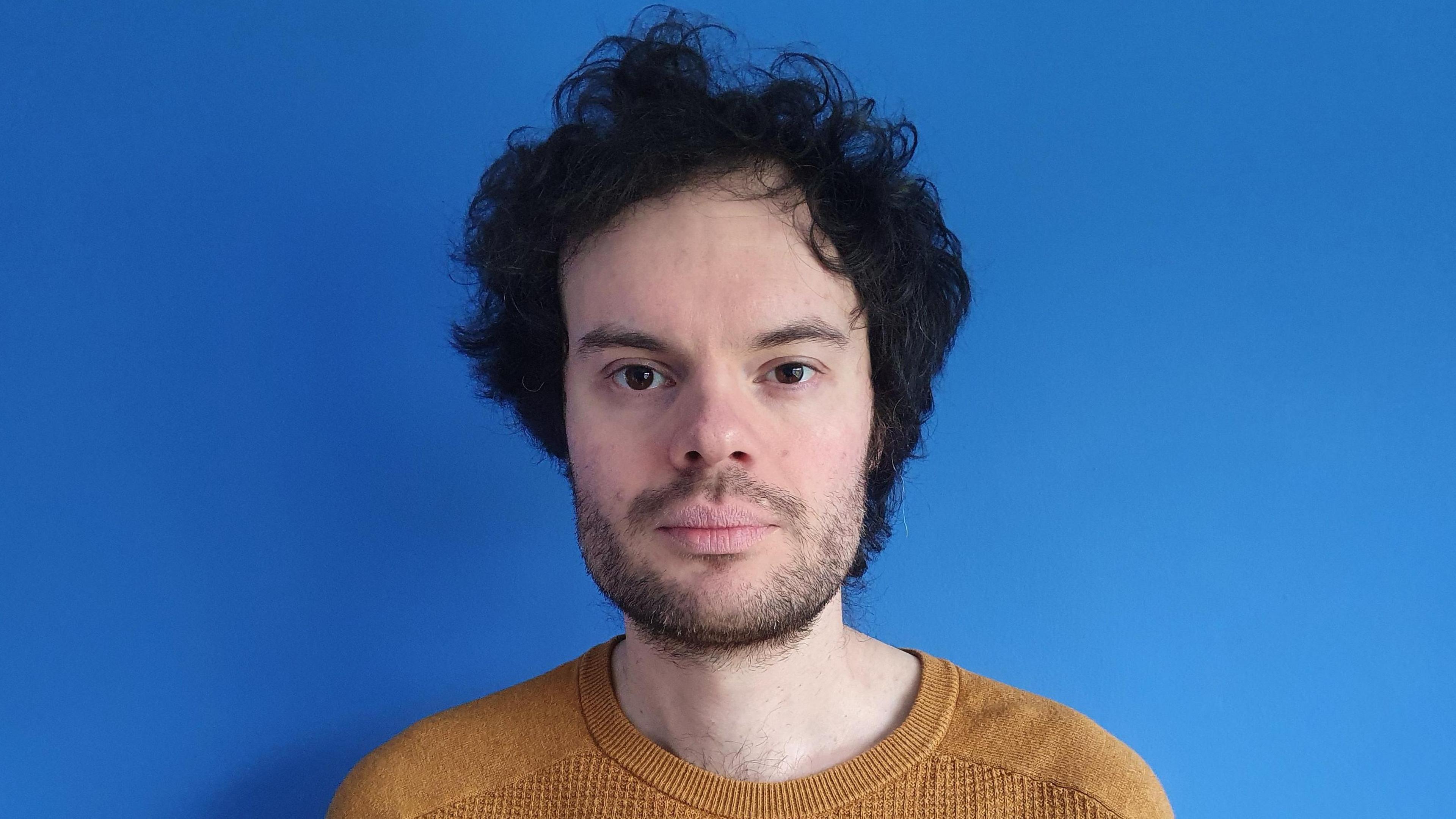 A man in a mustard yellow jumper with black stubble and thick black hair stands in front of a blue wall