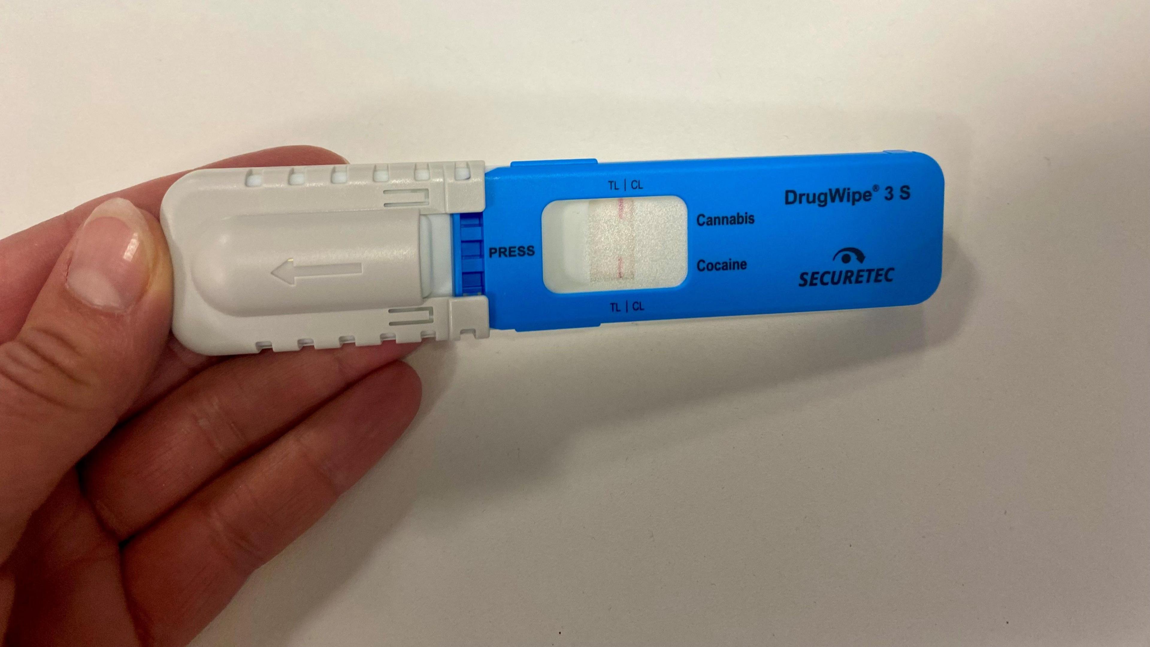 A DrugWipe test. It is blue and white and shows a reading for cannabis and cocaine, with a red stripe in a viewing window. It is being held in someone's hand.