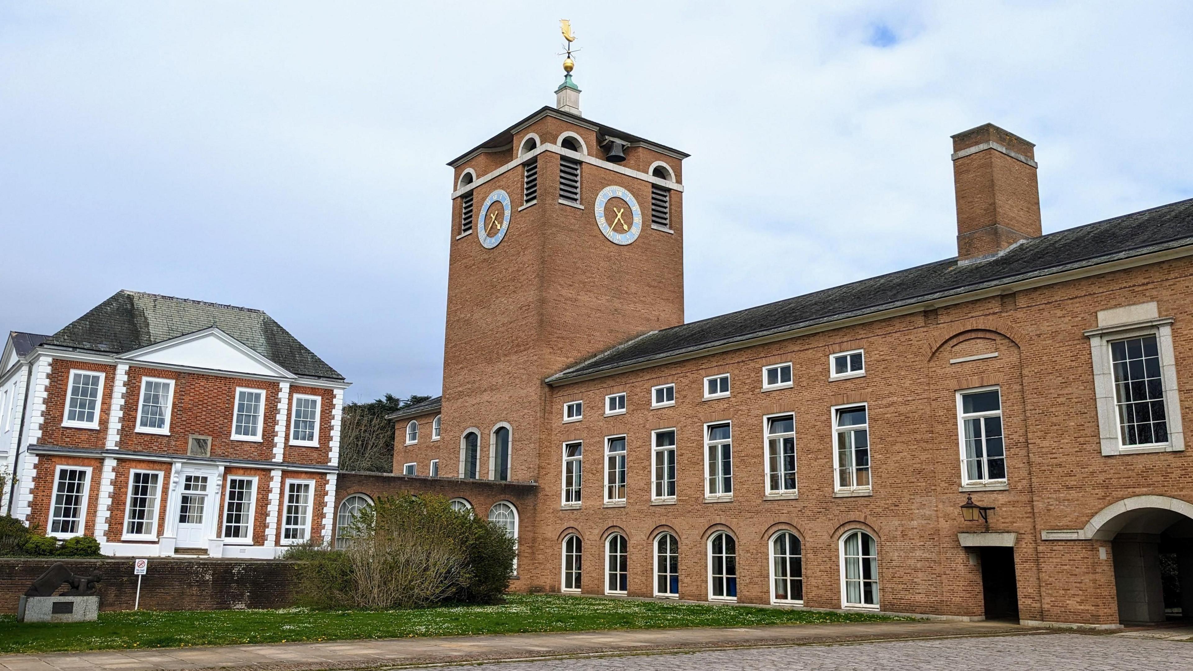 County Hall in Exeter, the base of Devon County Council. It consists of a large brown brick built building with a clock on the tower of one of the buildings. There is a weather vane on top of the tower.