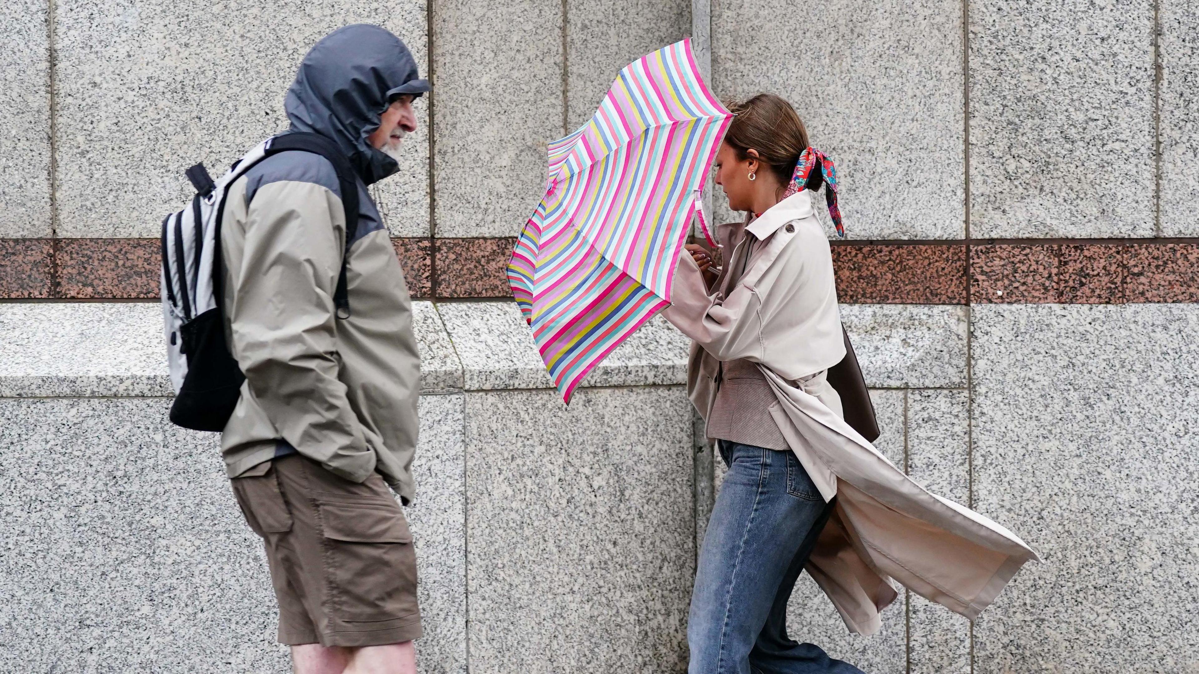 A man wears a raincoat with the hood up, over a pair of shorts, next to a woman shielding herself from the wind with a stripy pink umbrella, against a grey building background