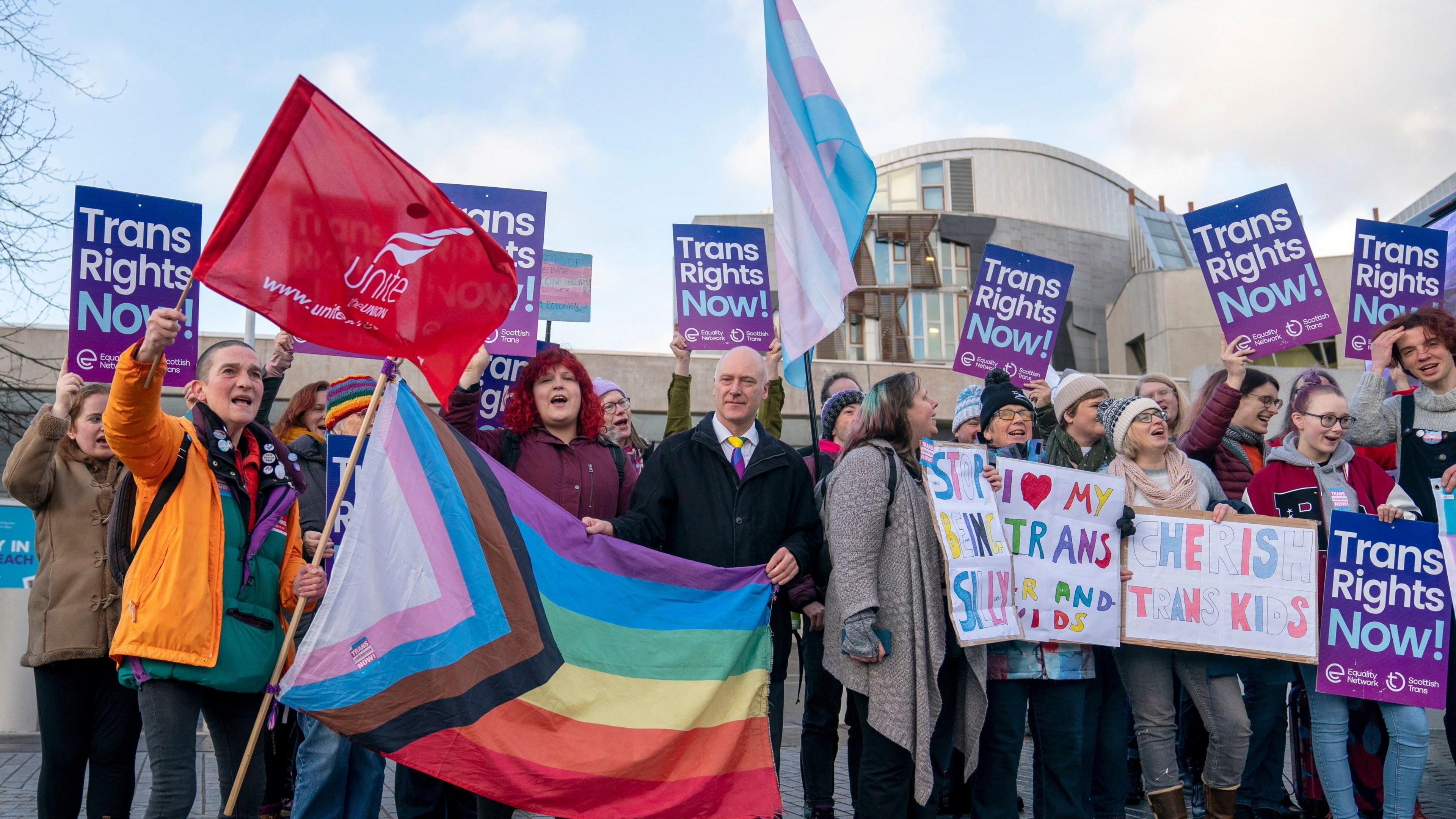 Trans rights campaigners outside the Scottish Parliament, holding up a flag in their colours, and placards reading "trans rights now" and "cherish trans kids"