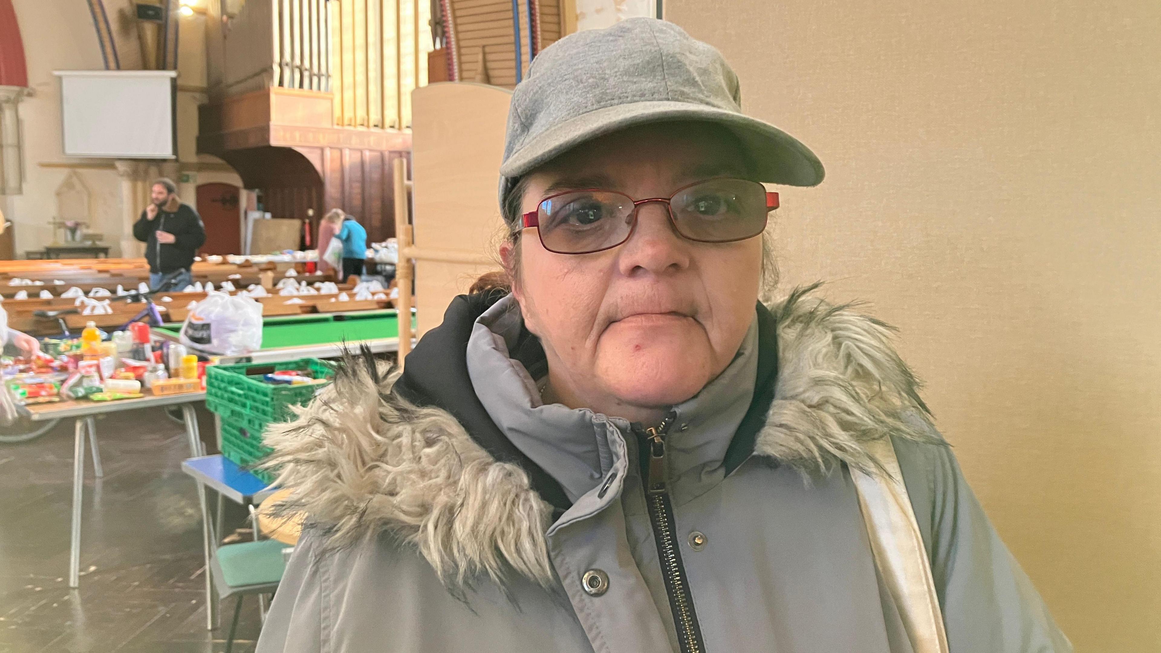 A woman wearing a grey winter coat, cap and red glasses looks into the camera. Food parcels can be seen in the background.