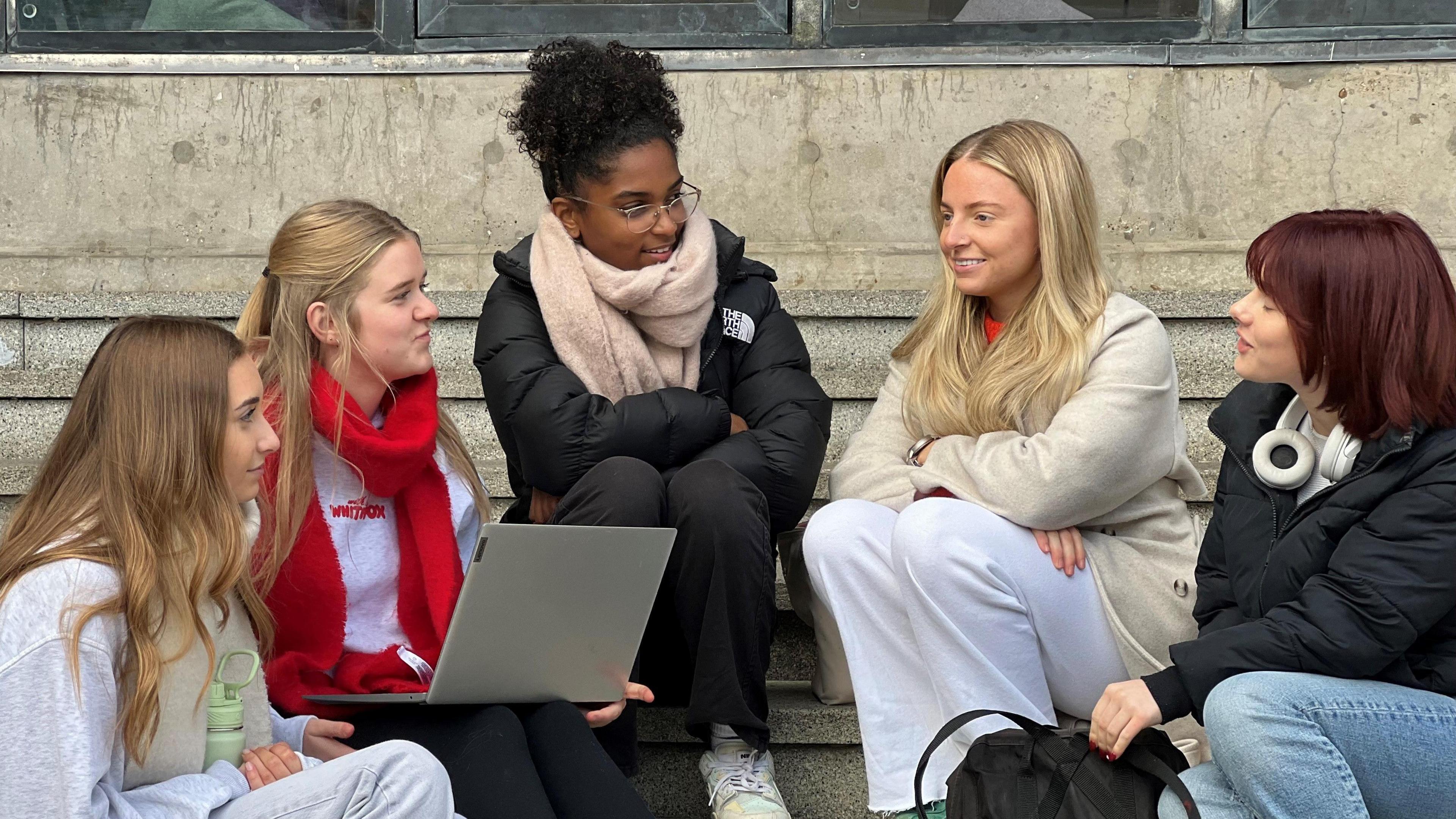 A group of students sit on steps on a university campus. They are chatting and laughing.