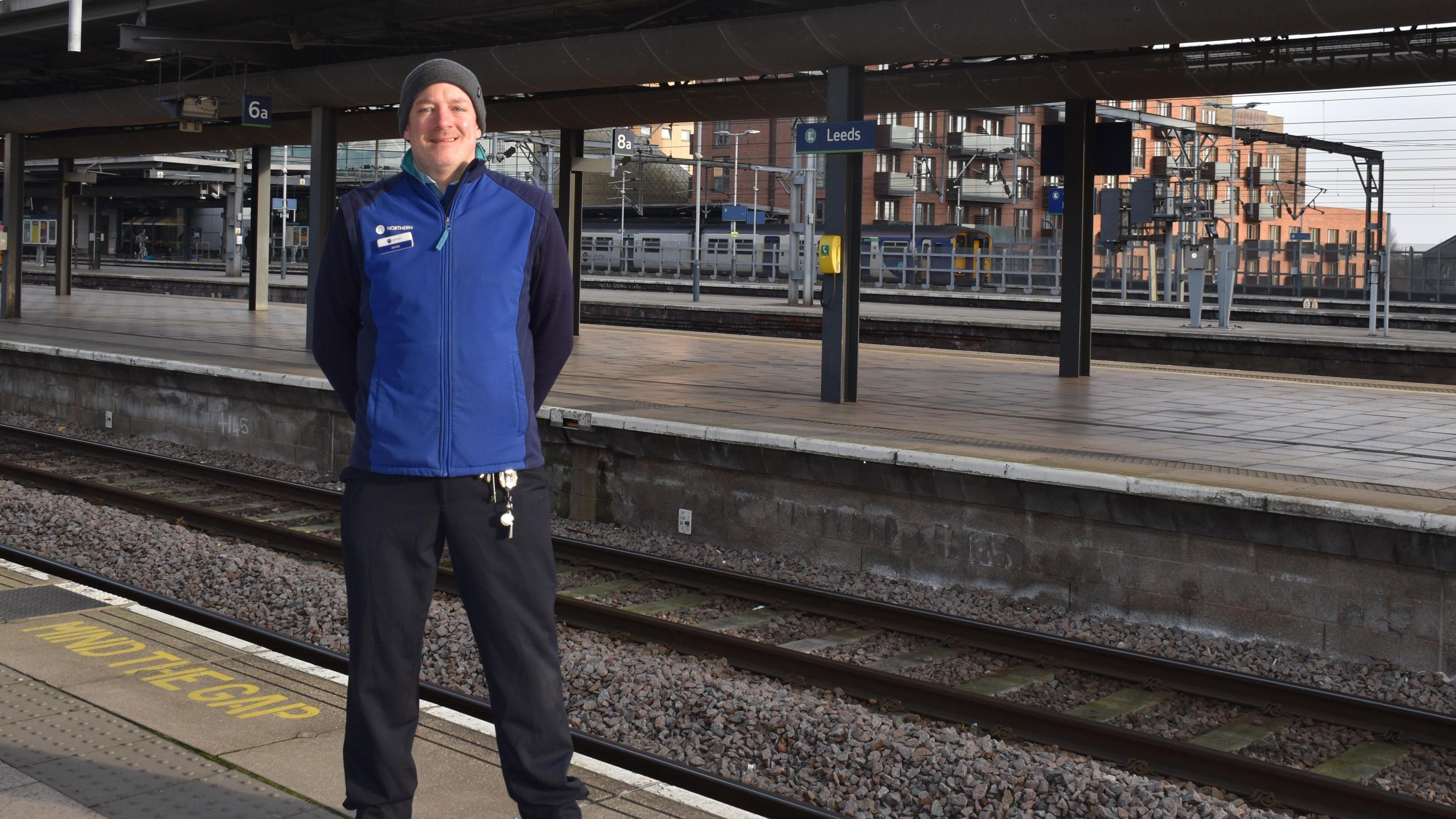 A train conductor wearing a blue uniform stands in the train station on the platform 