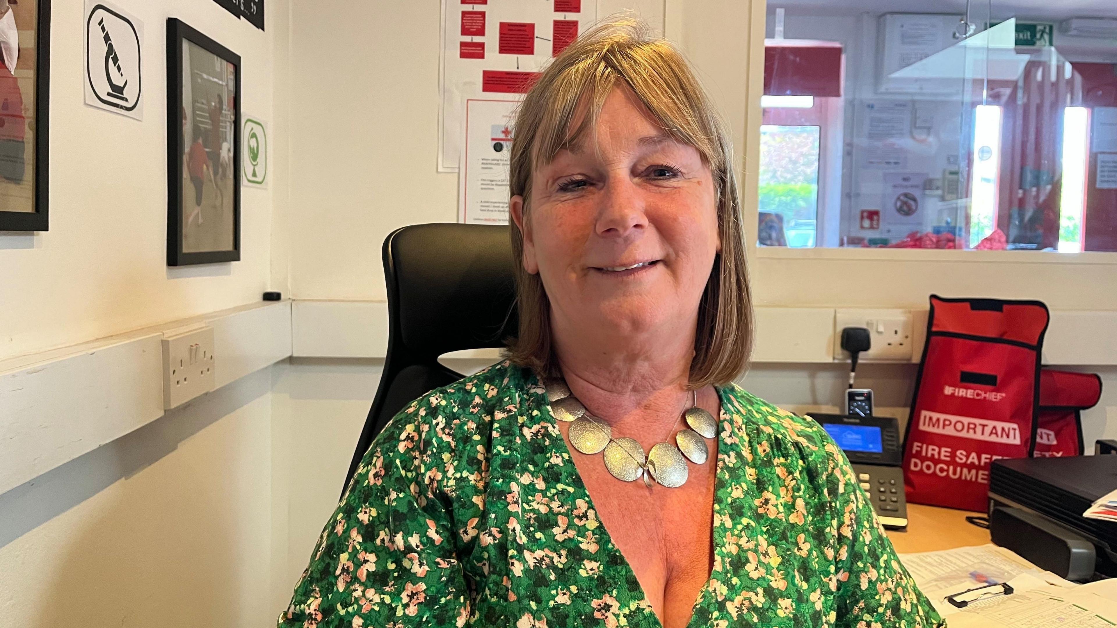 Carolynn Southcombe smiling at the camera in her office. She has brown shoulder-length hair and is wearing a green flowery top.