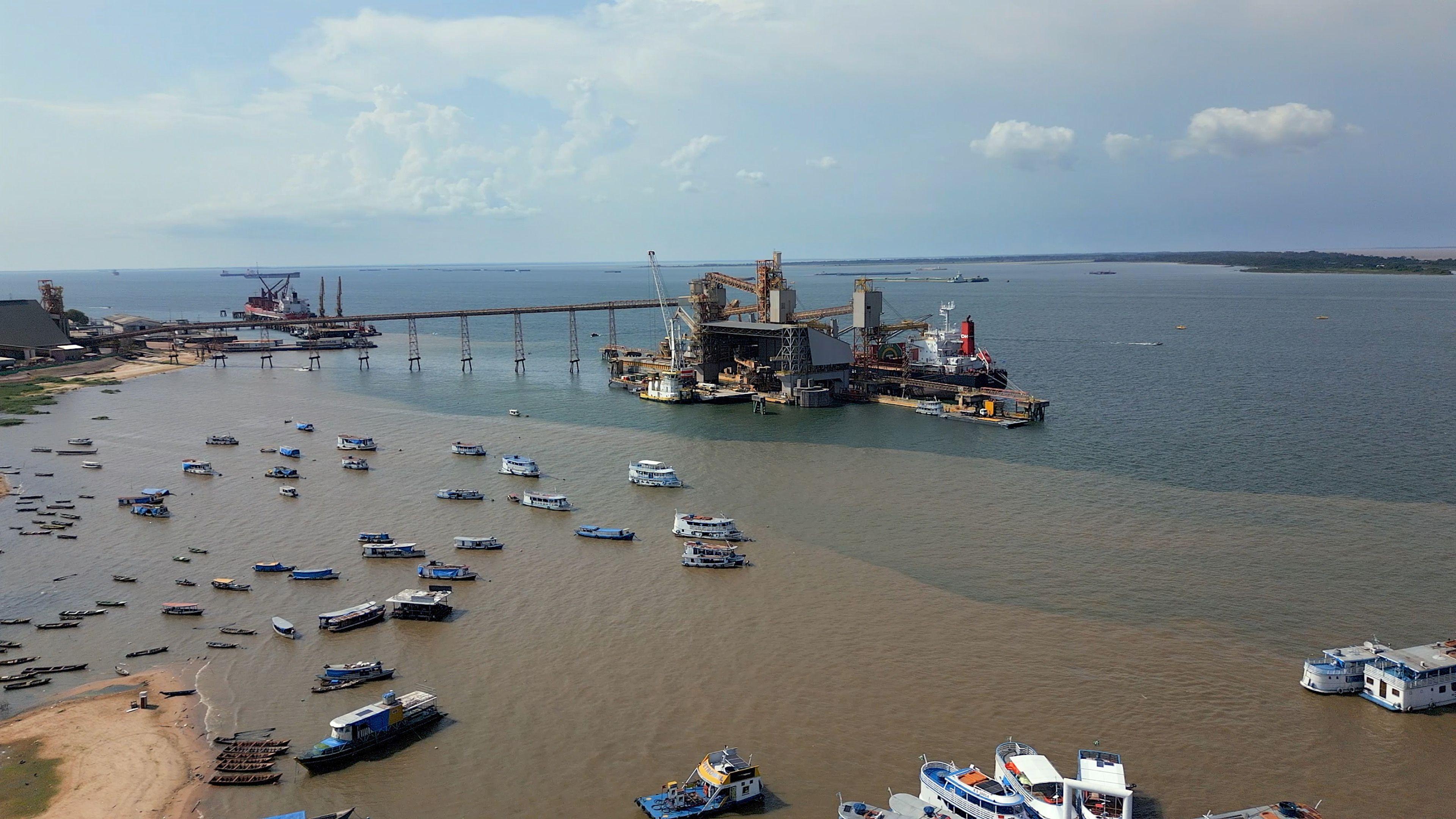 A wide view of a port at the mouth of the Tajapos River in the Amazon river. You see a terminal and an industrial complex which is connected by a bridge. A number of ships and boats are in the water which looks brown close to the shore and blue away from the shore.