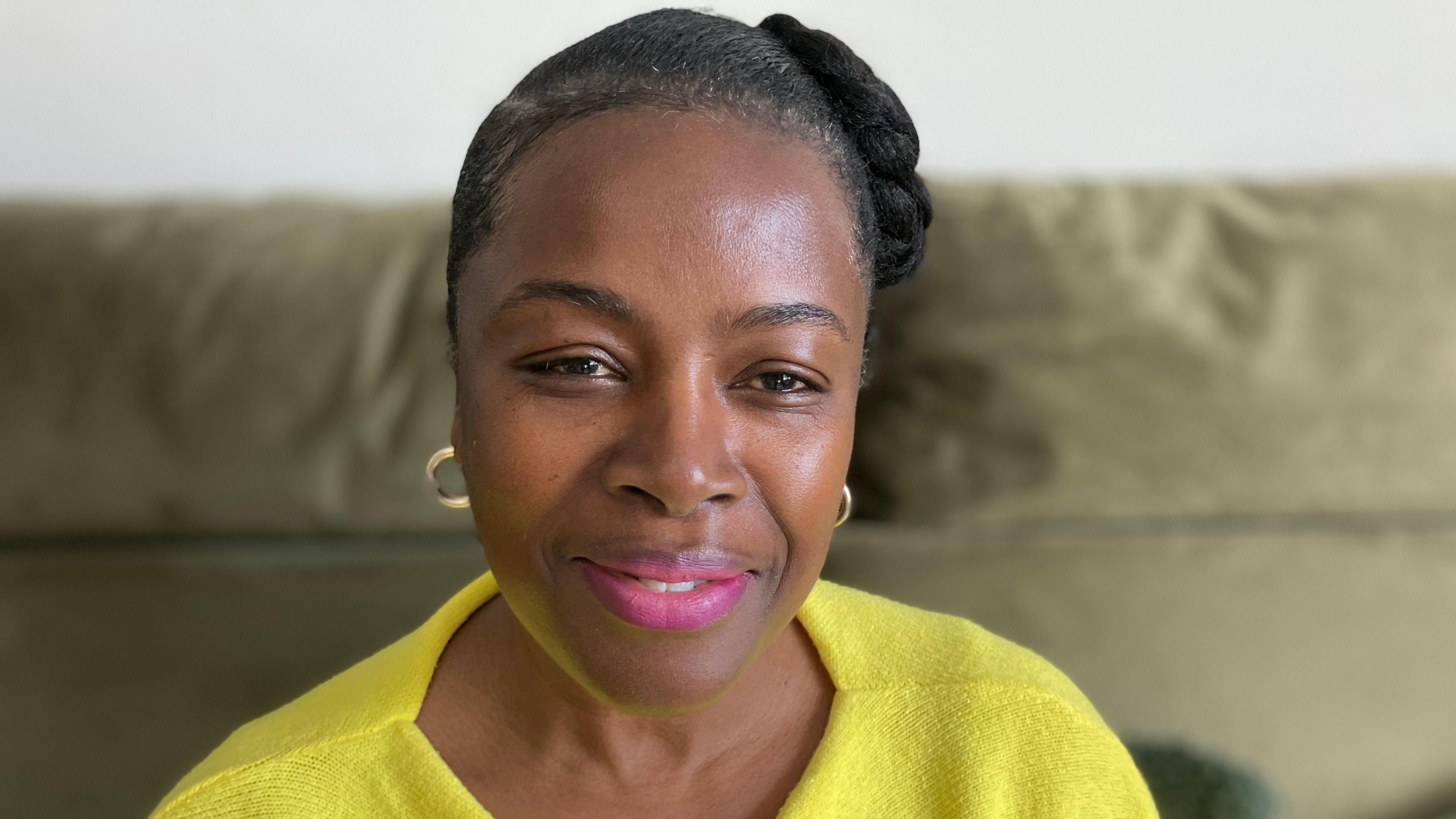 Sandra Smith-Brown is smiling and sitting down on her sofa in her house. She has black hair pinned back and is wearing a bright yellow jumper.