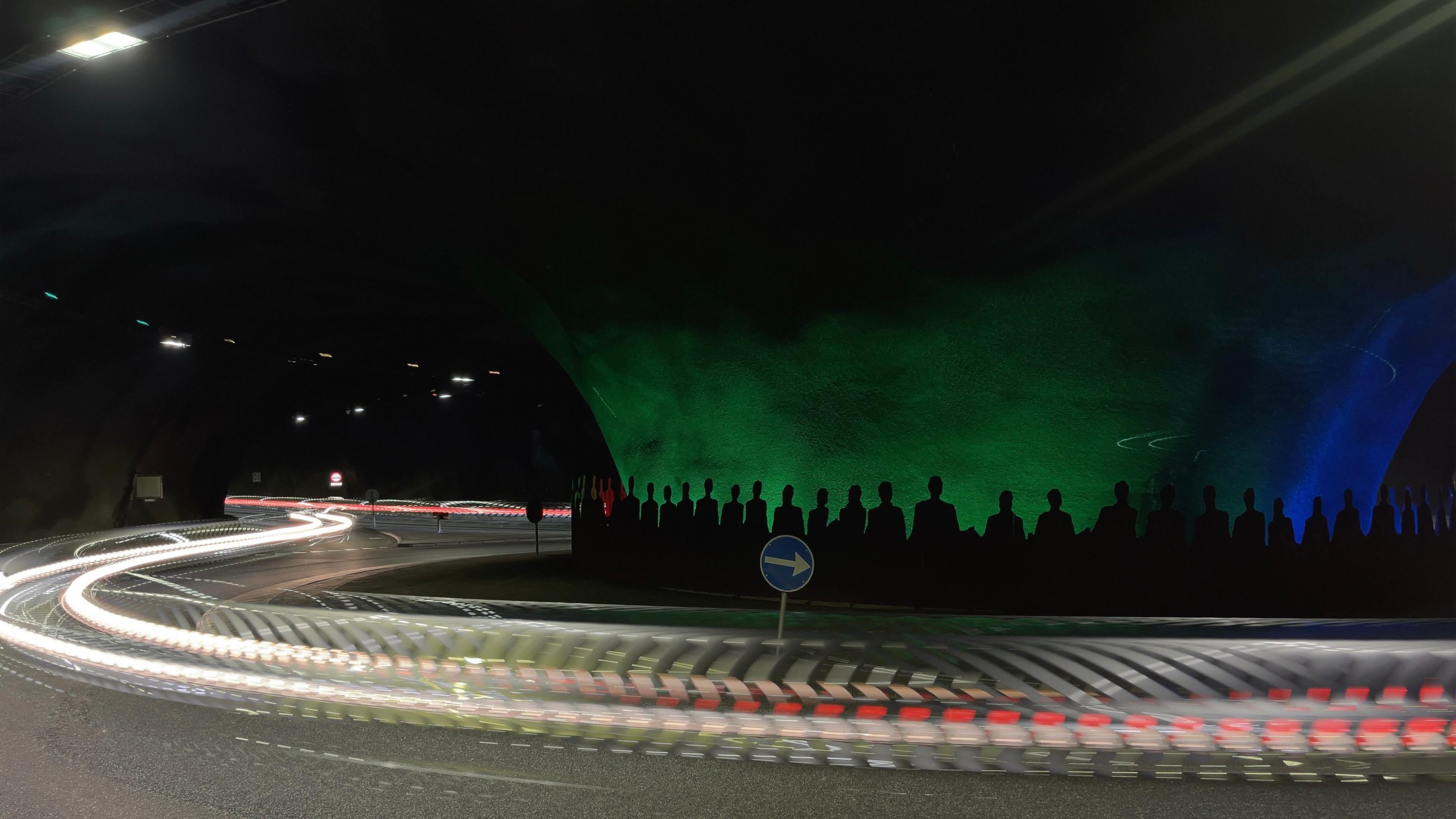 Photograph of vehicles streaking past a roundabout in a tunnel. The tunnel has a green/ blue backdrop and black silhouettes of figures on it.