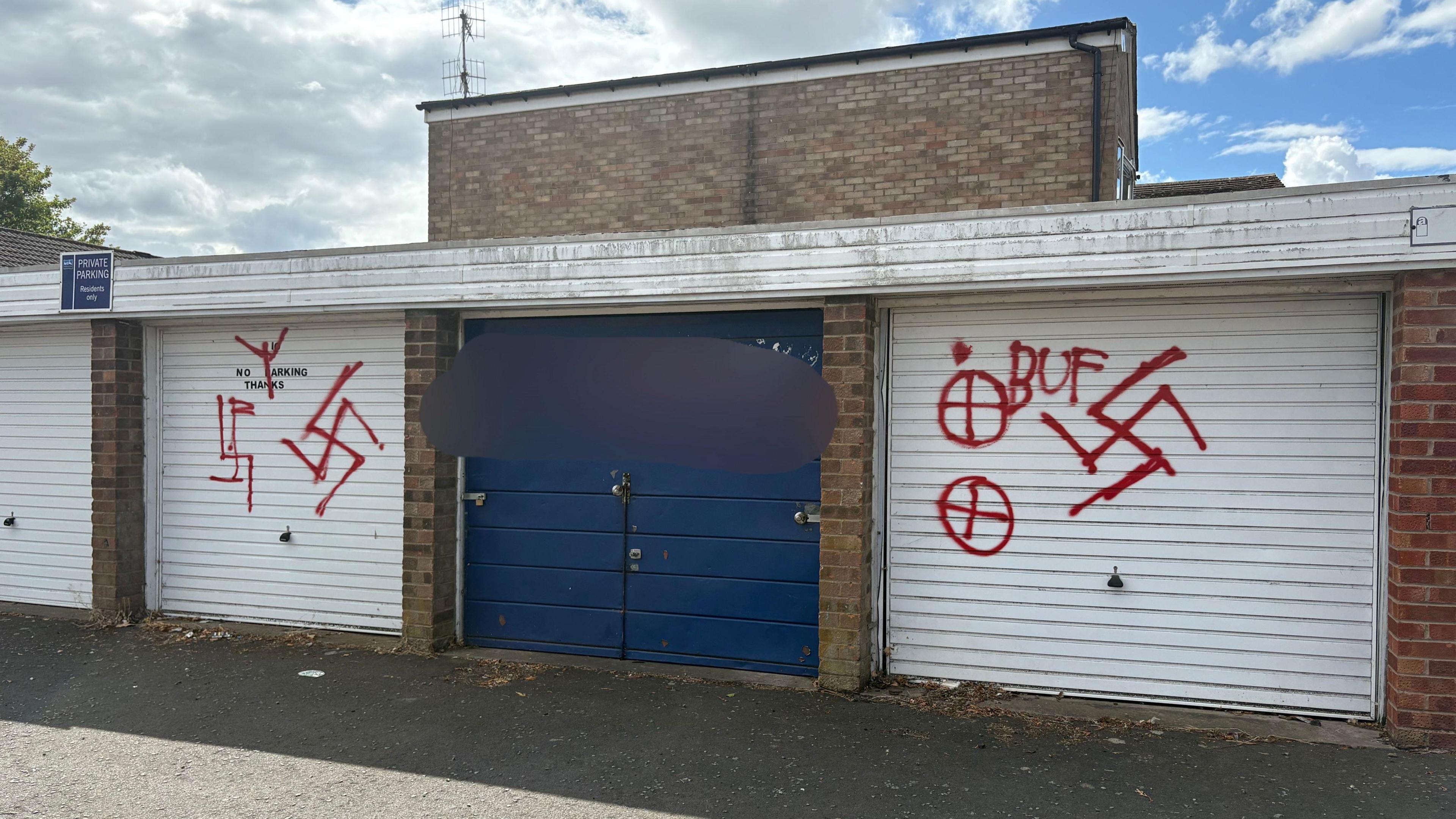 Three garage doors, two white and one blue, in a garage building. The white doors, either side of the blue one, have red graffiti including swastikas, crosses in circles and "BUF". The graffiti on the blue door has been blurred.