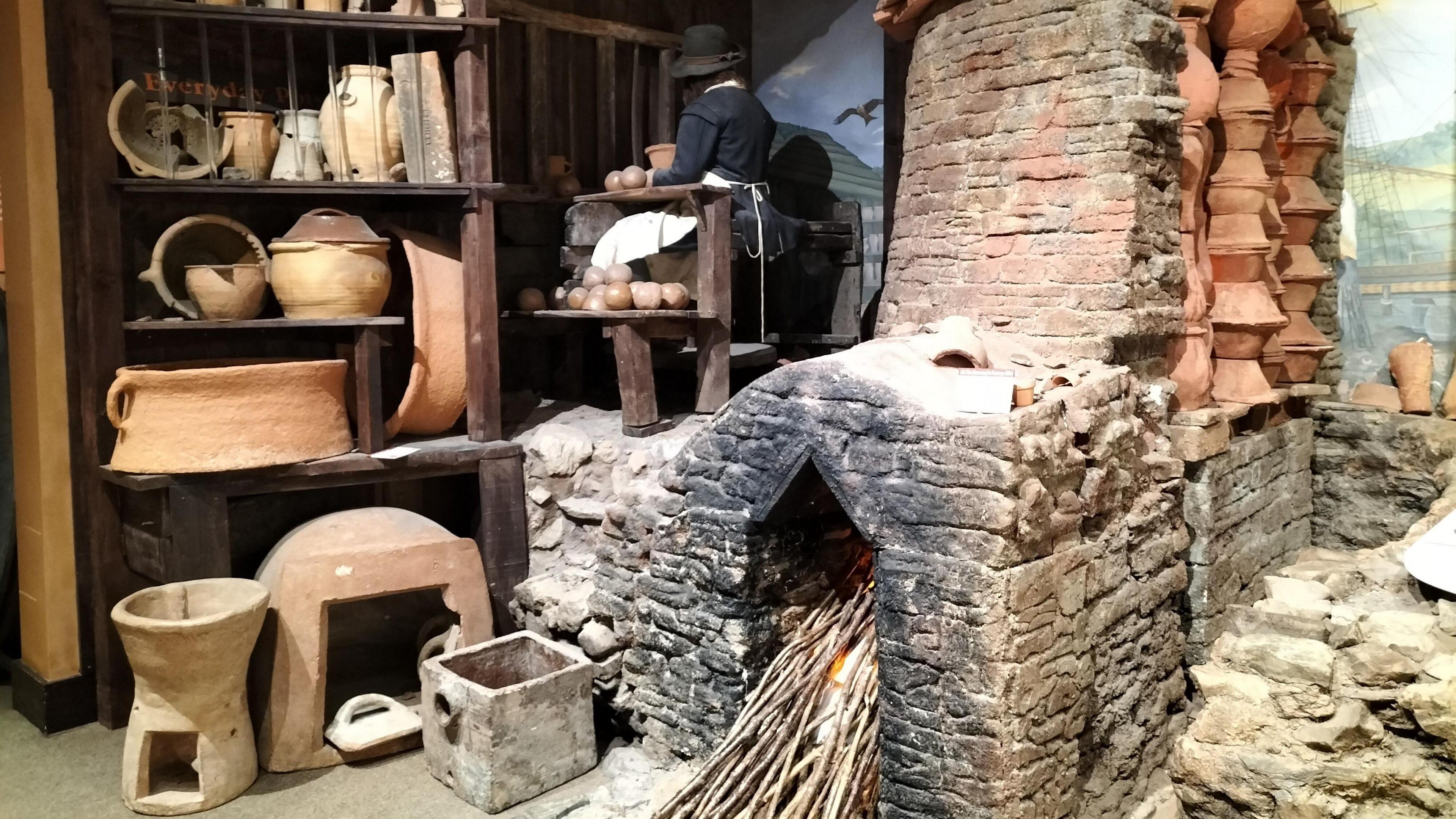 A room full of old pots and a large fire place in a kiln. There is wood stacked inside it and a fire taking place.