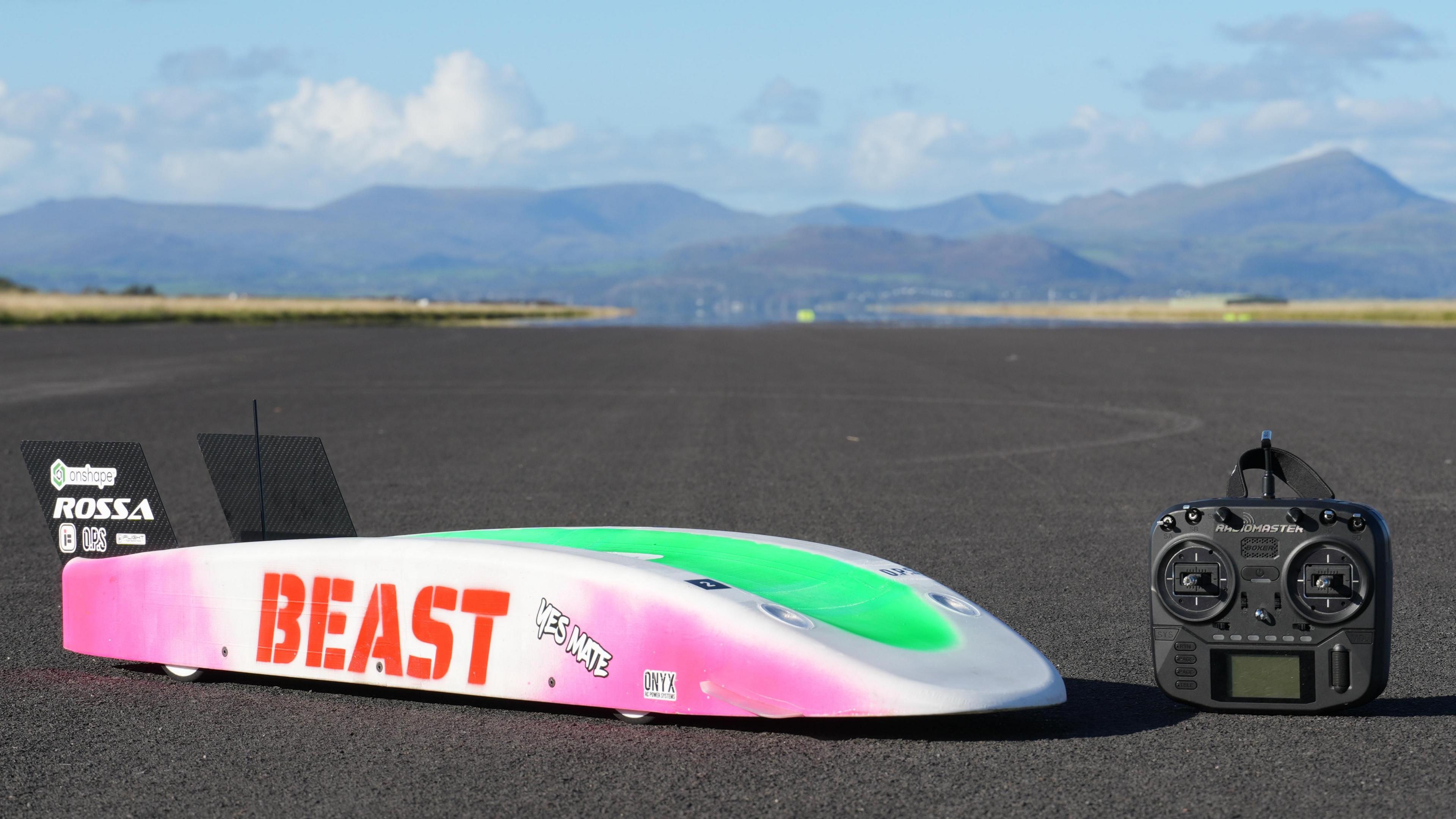 The image shows 'The Beast' remote control car on the left and a remote controller unit on the right. Beyond them is a smooth road surface leading to the horizon where there are hills against a blue sky