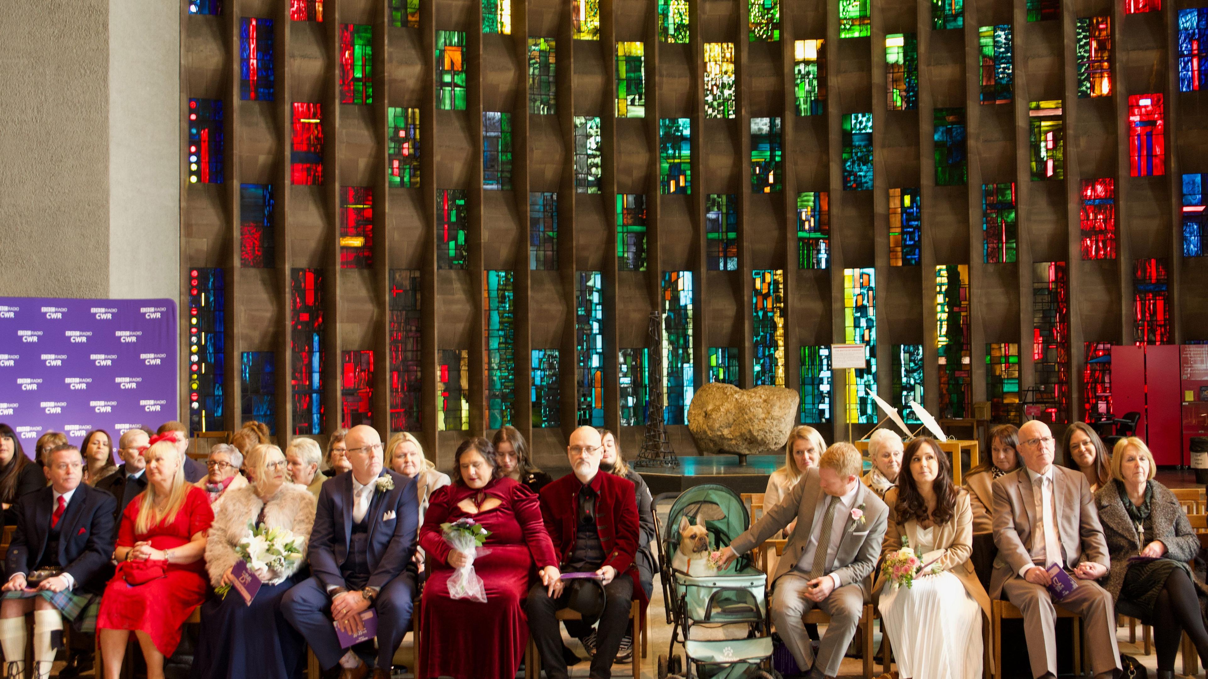 A stained glass window at the cathedral with two rows of people dressed in wedding outfits in front