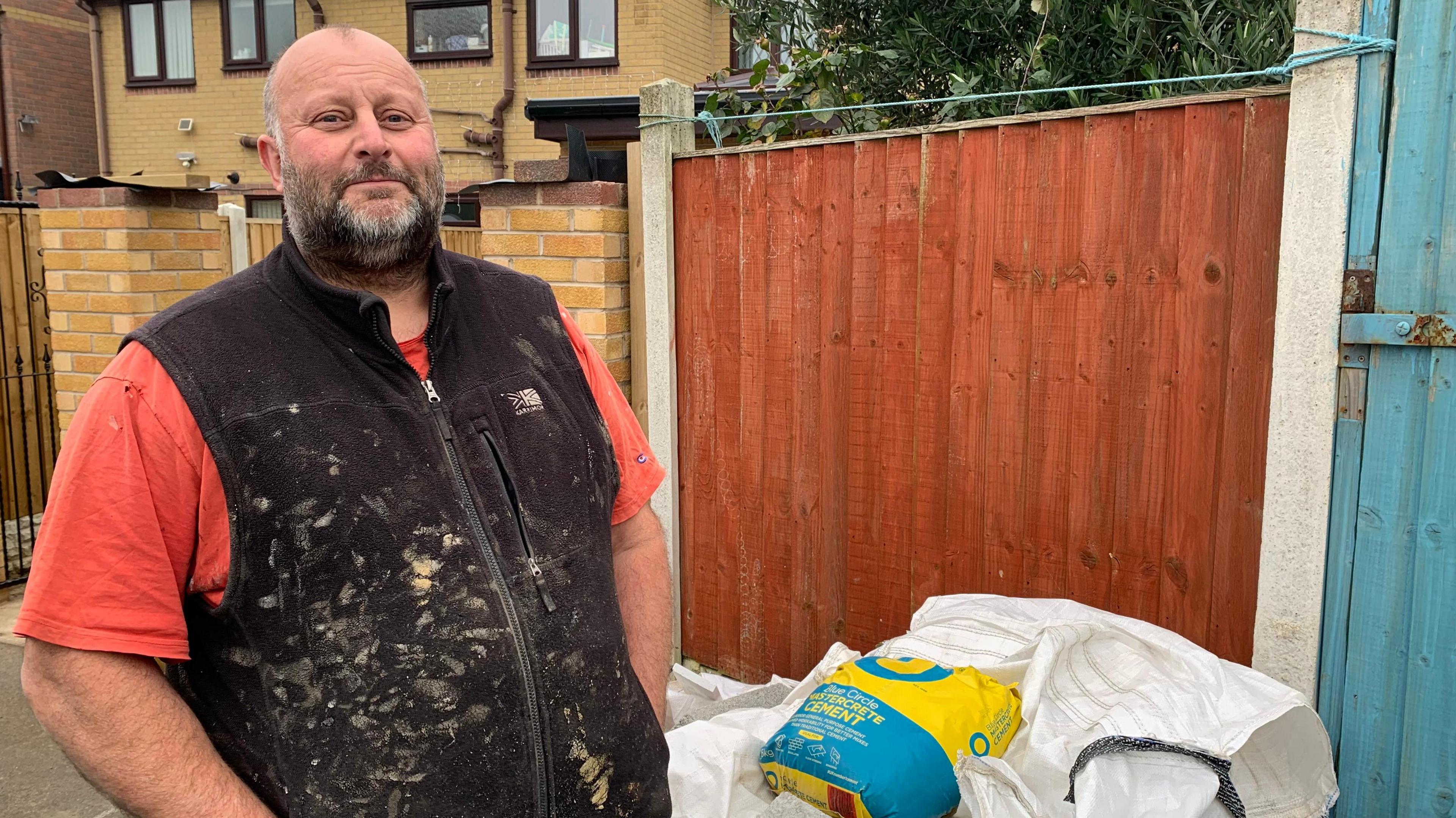 Simon Robinson stands outside his daughter's home in Catcliffe, which he is repairing after the flood. He has a short salt and pepper beard and moustache and is bald. He is a wearing a dirty black vest and orange t-shirt. There is a large white canvas on the floor and a bag of cement.