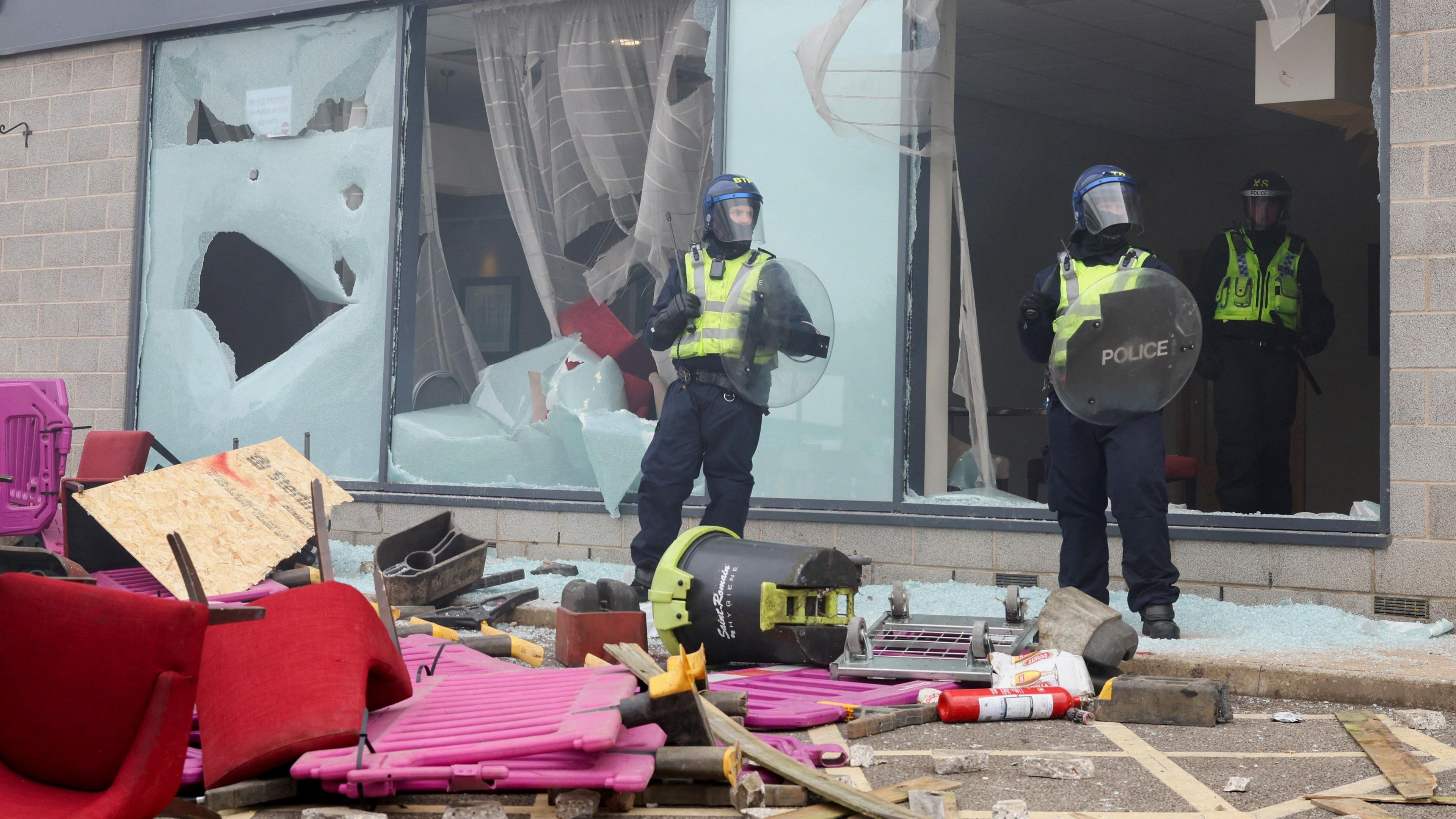 Three police officers in riot gear stand amid piles of rubble outside a hotel with broken windows