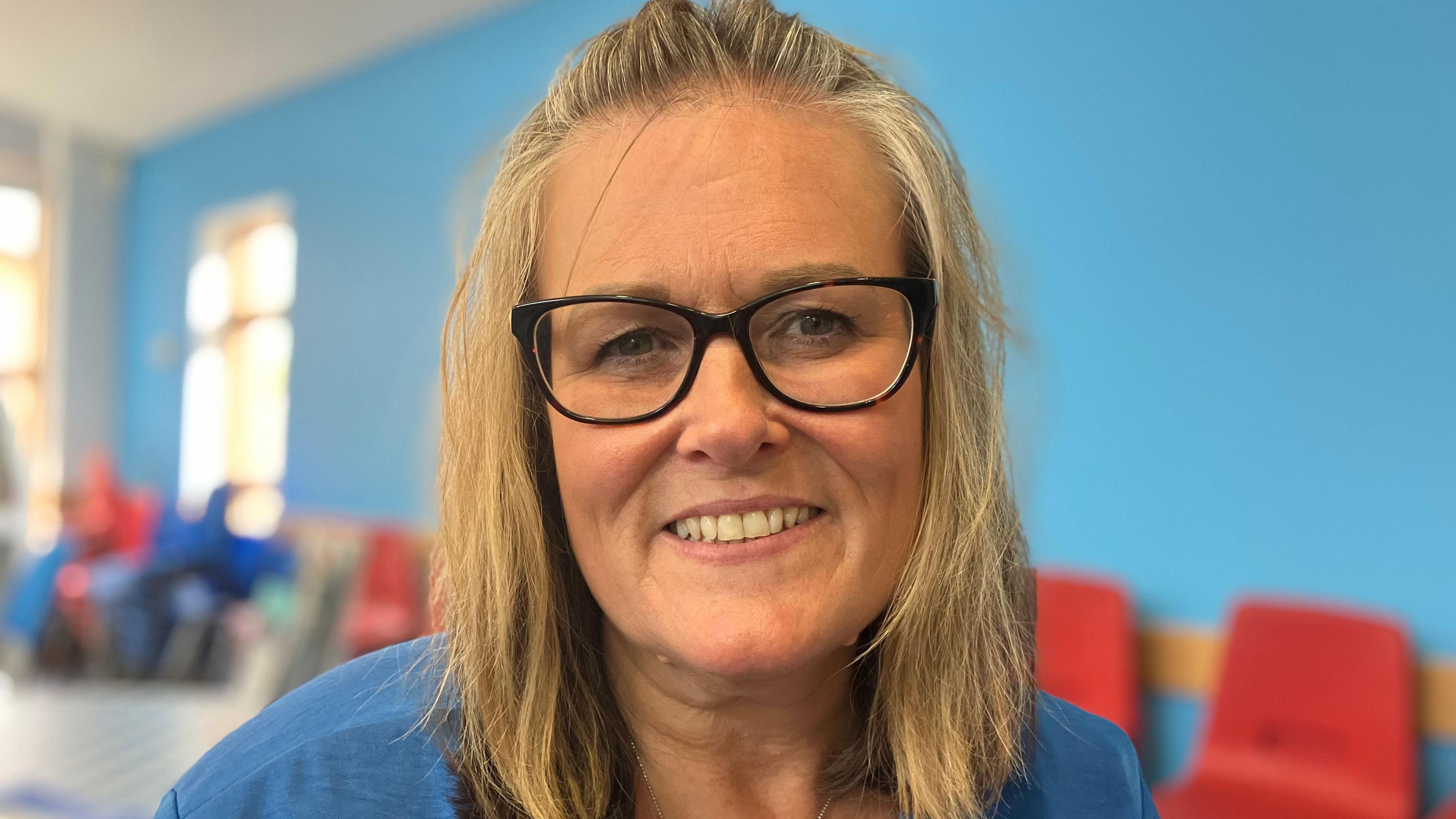 A blonde woman with black glasses with a blue wall and a row of plastic chairs in soft focus behind her.