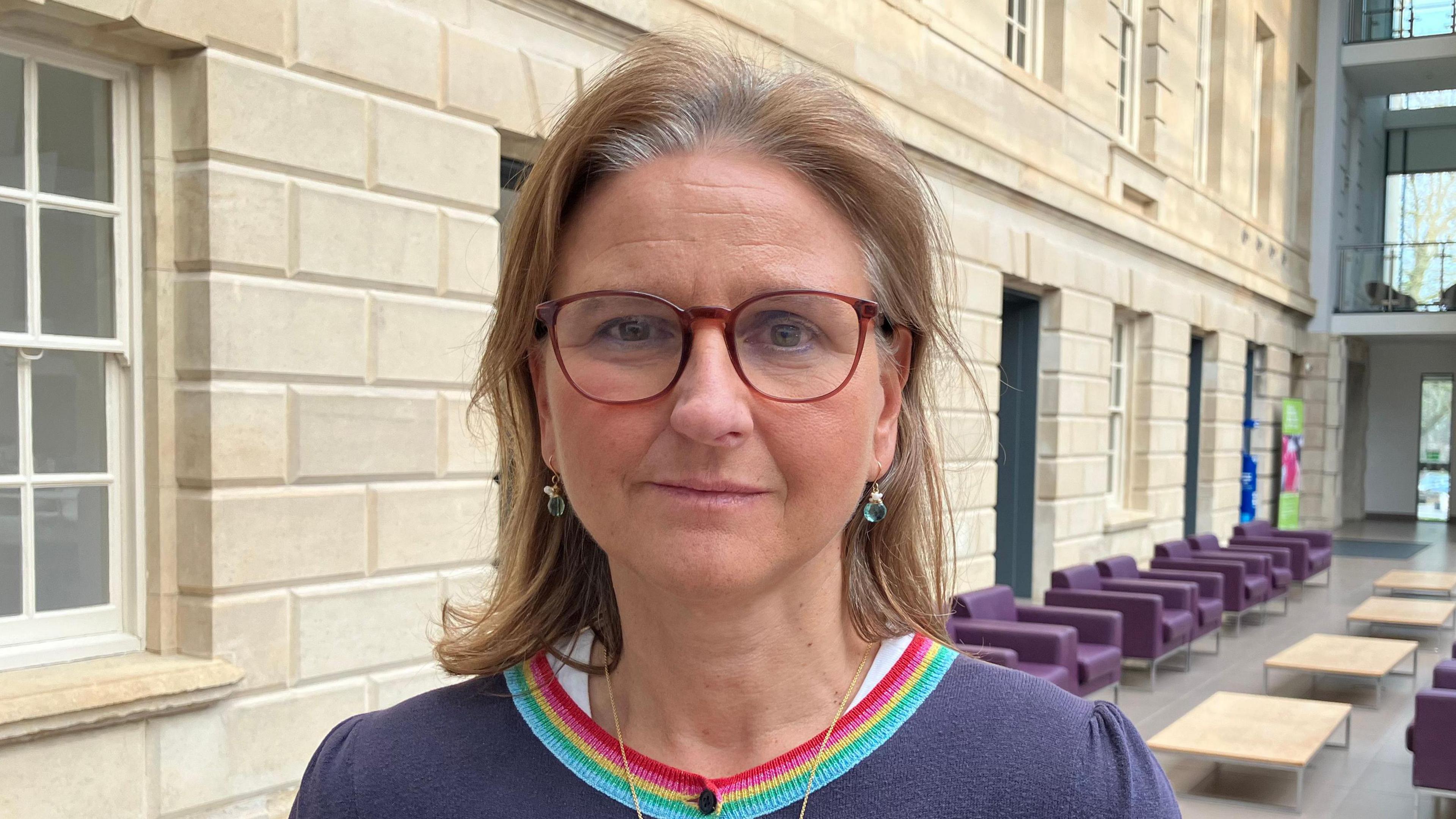 Laura Mayes, looking at the camera. She has brown glasses on and long brown hair, and is photographed inside an office building
