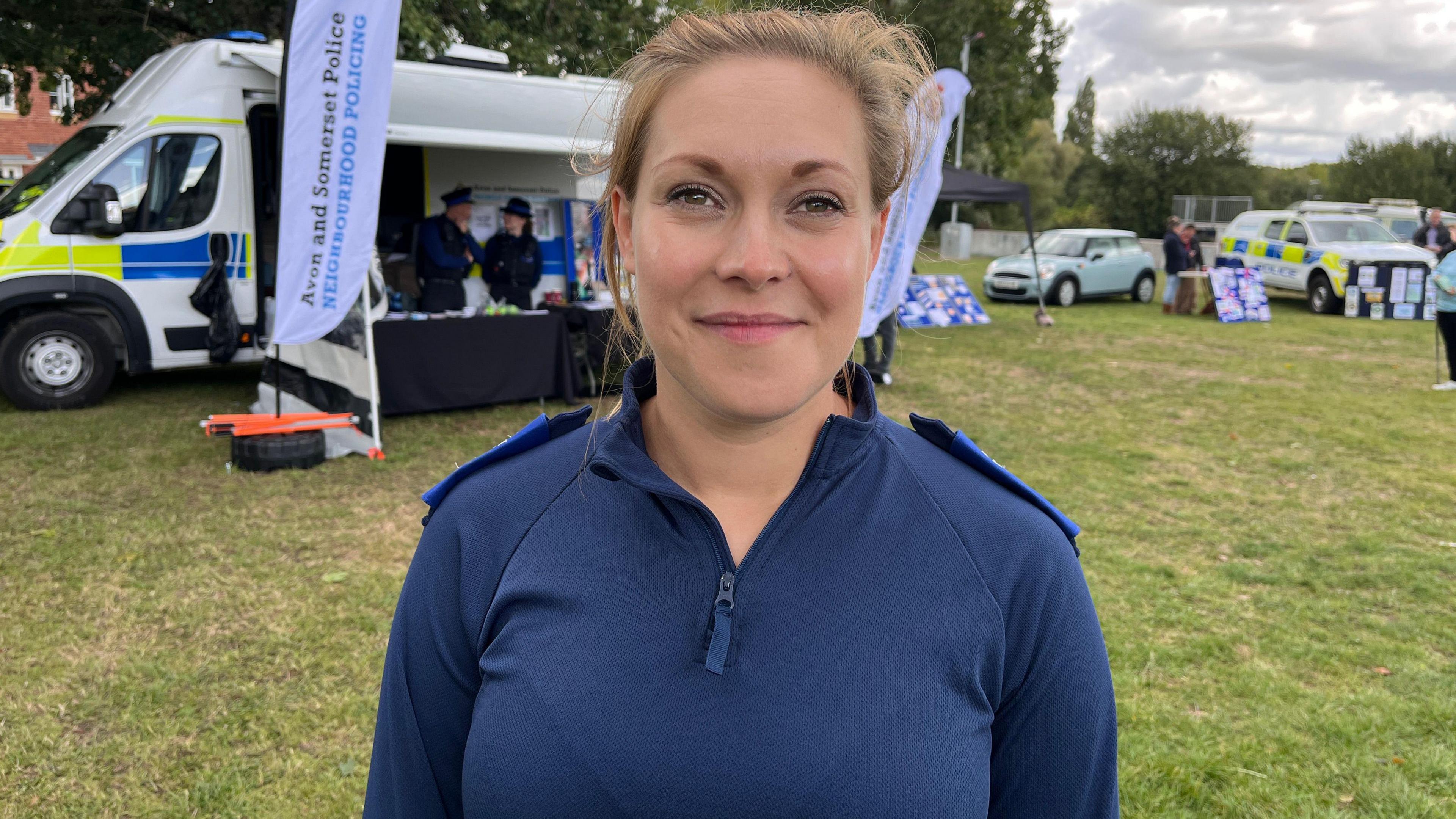 PCSO Bea Gane is looking at the camera, she is smiling and wearing a blue three-quarter zipped jacket with police epiliets on her shoulder. Behind her is a police van with flags flying outside it that say Avon and Somerset Police, Neighbourhood Policing.