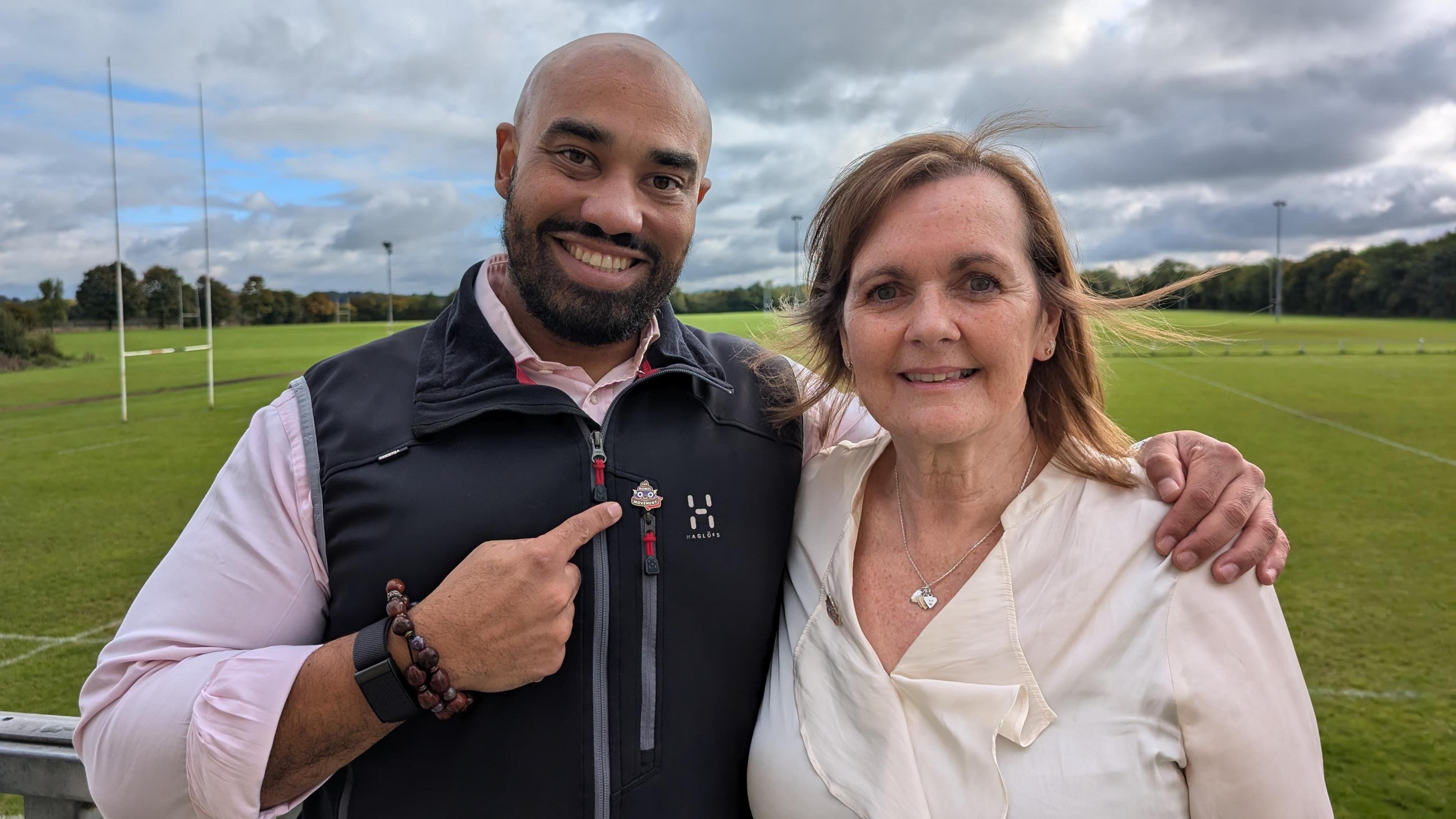 Ram Patten has his arm around Jo Millard. Ram is pointing at Jo. They are both smiling. They are standing beside a rugby field.