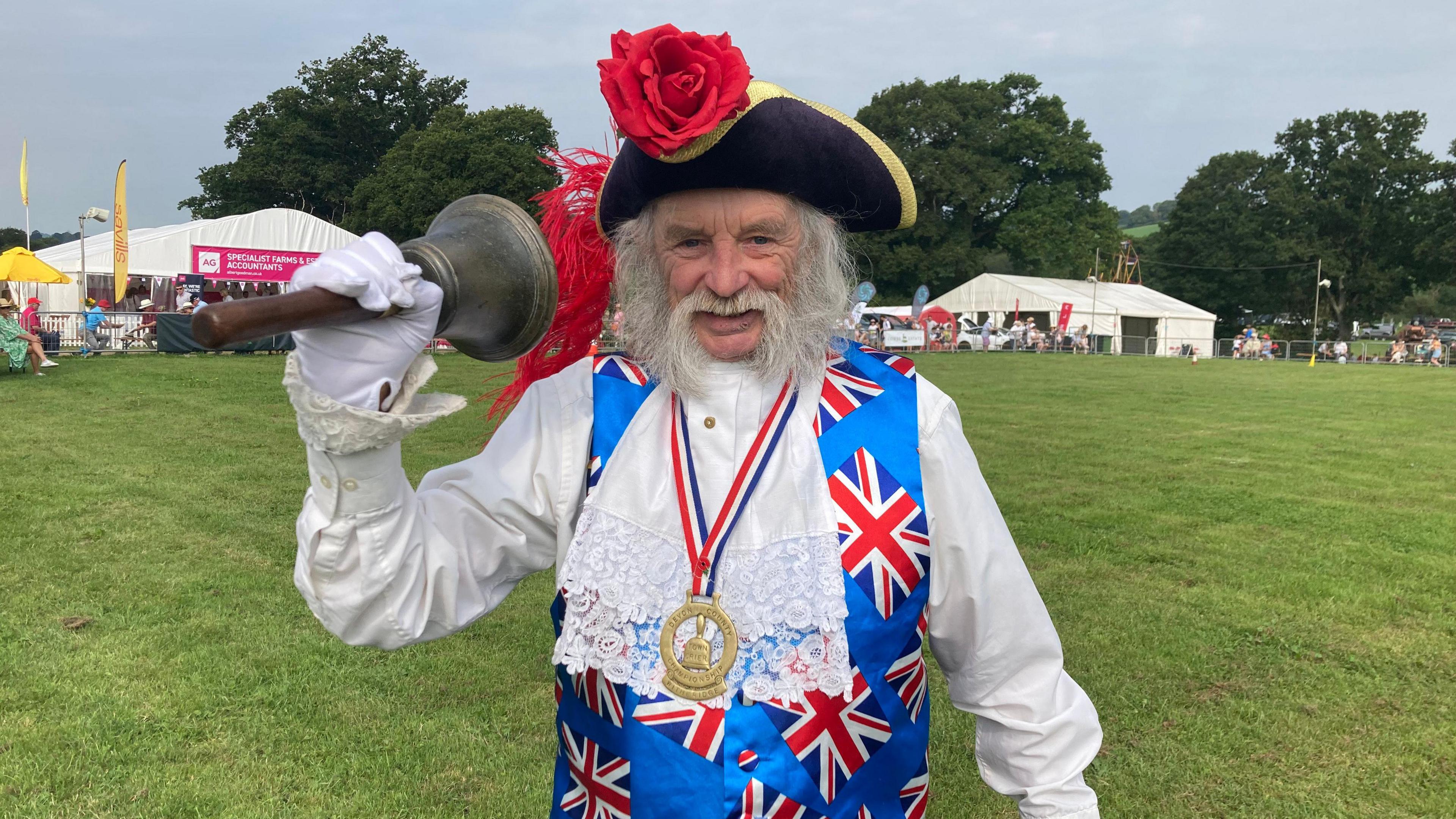 Dave Retter smiles at the camera wearing a black and gold tricorn hat with a red rose on it, a frilled sleeve white shirt, blue Union Jack waistcoat, medal and ruff. He has white hair and mustach and is ringing a large bell with a wooden handle