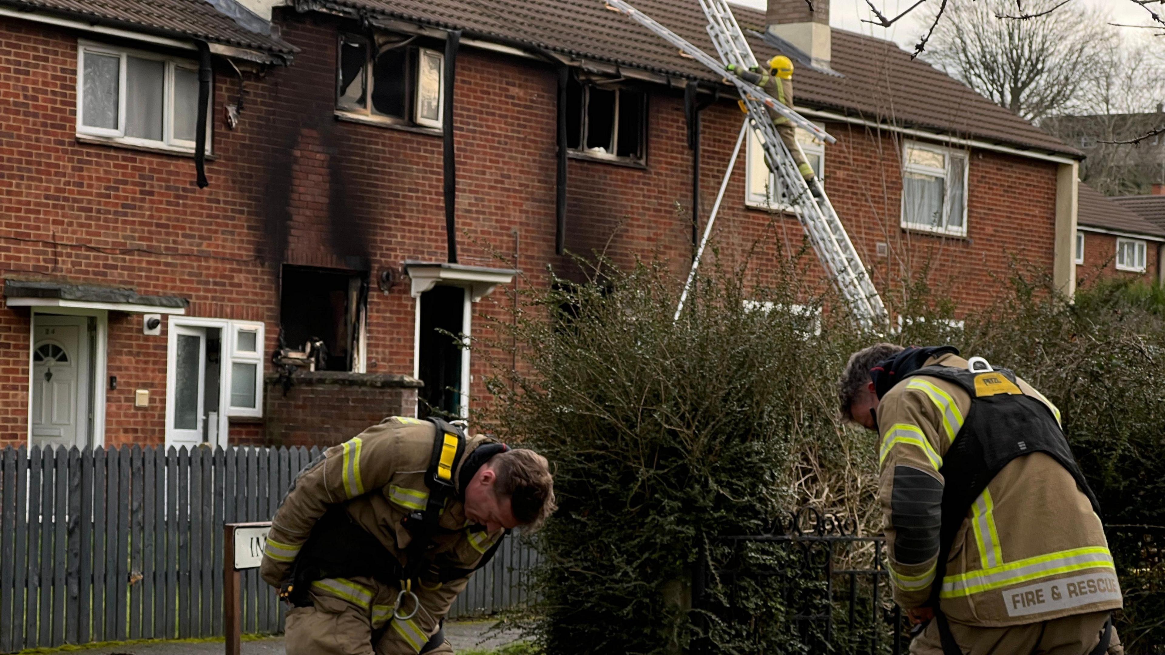 Two fire officers at the scene in Imber Walk in Swindon. It is a residential area with a number of red brick terraced properties. There is significant damage to the facade of the house, with black smoke damage going up the wall. A third firefighter is using a ladder to access the roof.