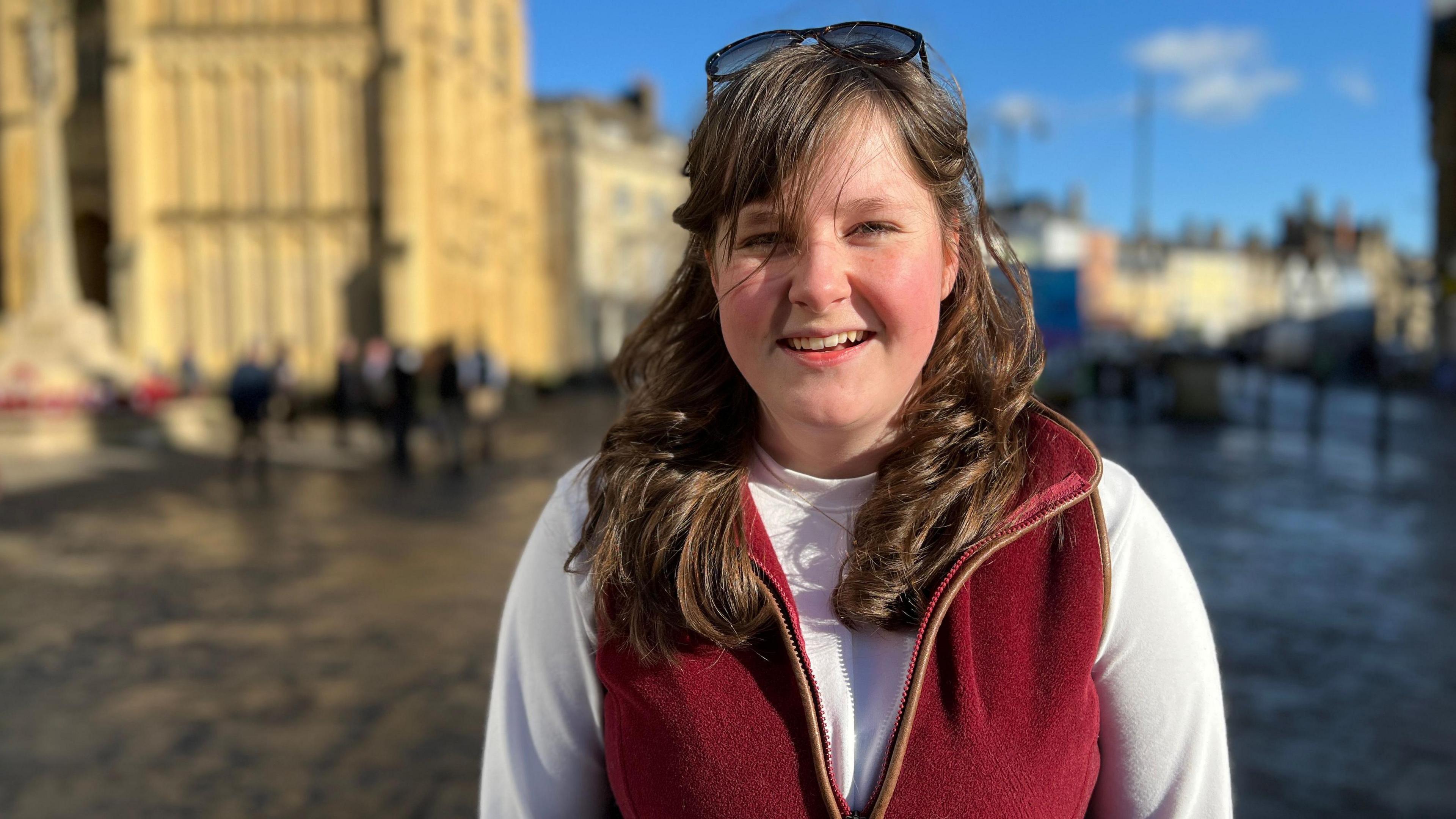 A young girl stands in Cirencester town centre, a church behind her, smiling on a sunny winter's day. She is wearing a white long-sleeved top and a red fleece gilet, with glasses on top of her brown hair, which is past her shoulders.