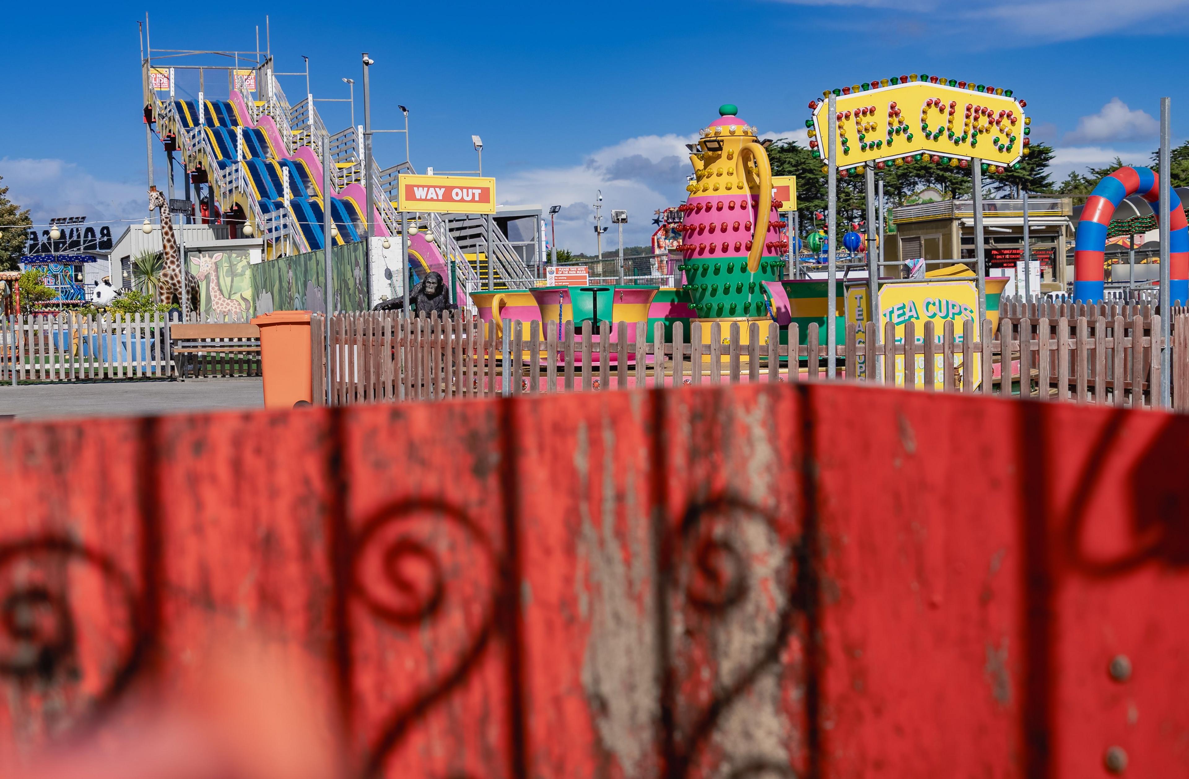 A red fence obscures the fair. The tea cups ride and slides can be seen behind. 