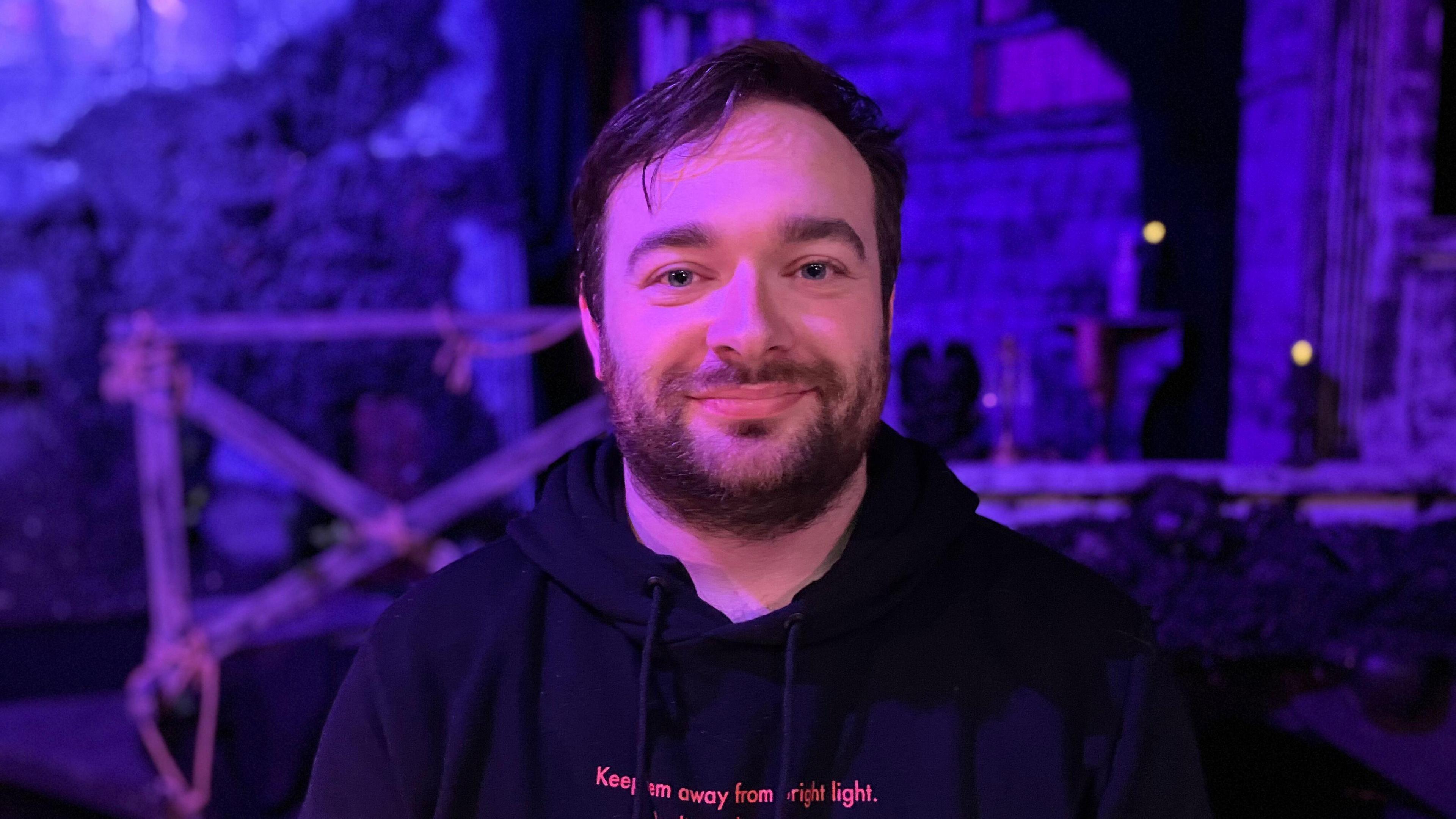 A man with black hair and short beard, he has blue eyes and wearing a black hoodie. He is looking at the camera with a smile on his face.