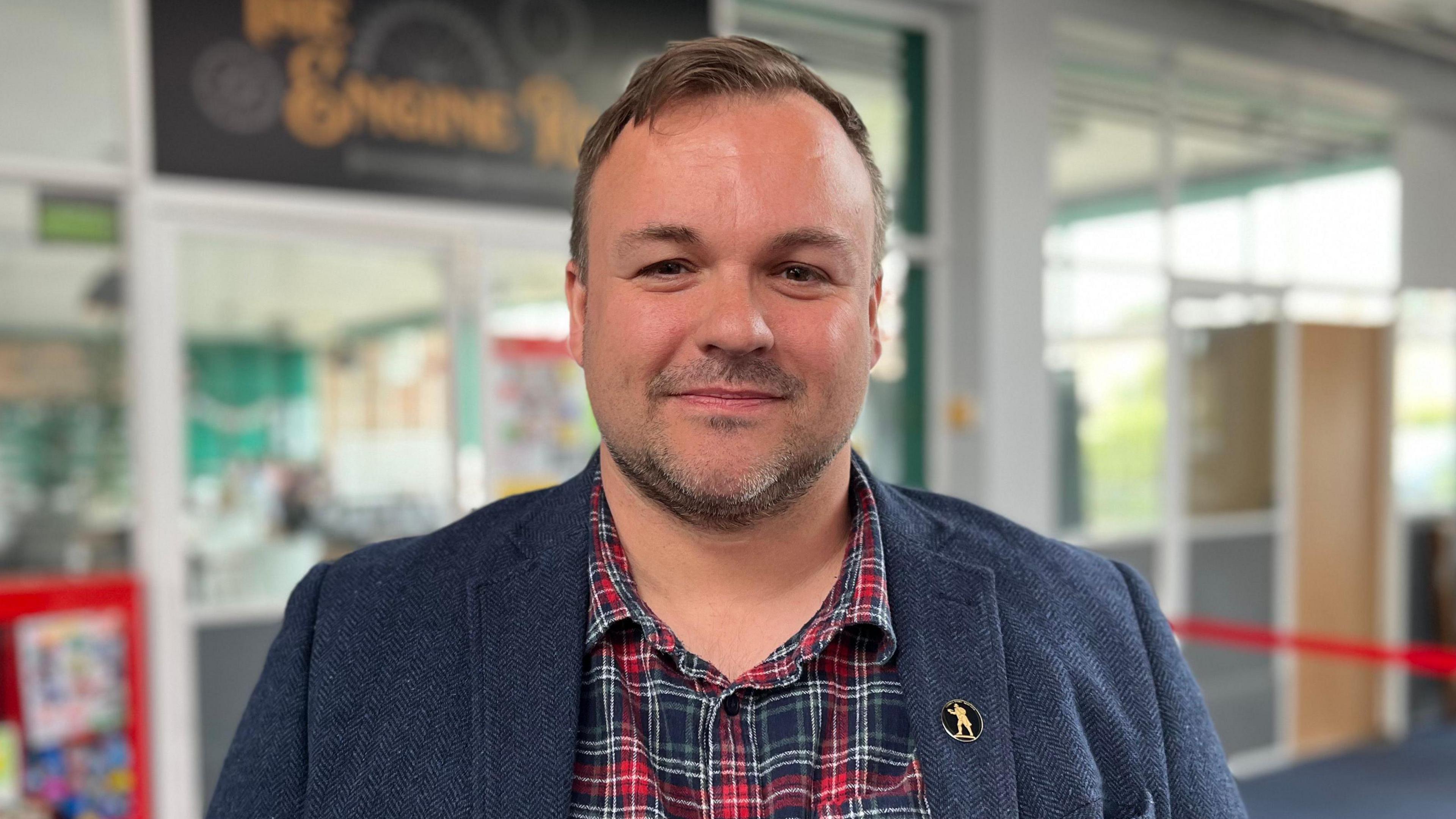 Terry Jermy is standing in the middle of the picture staring at the camera. He is wearing a navy blue coat, with a red and blue tartan shirt underneath. He has brown hair and a brown beard, and is smiling at the camera.