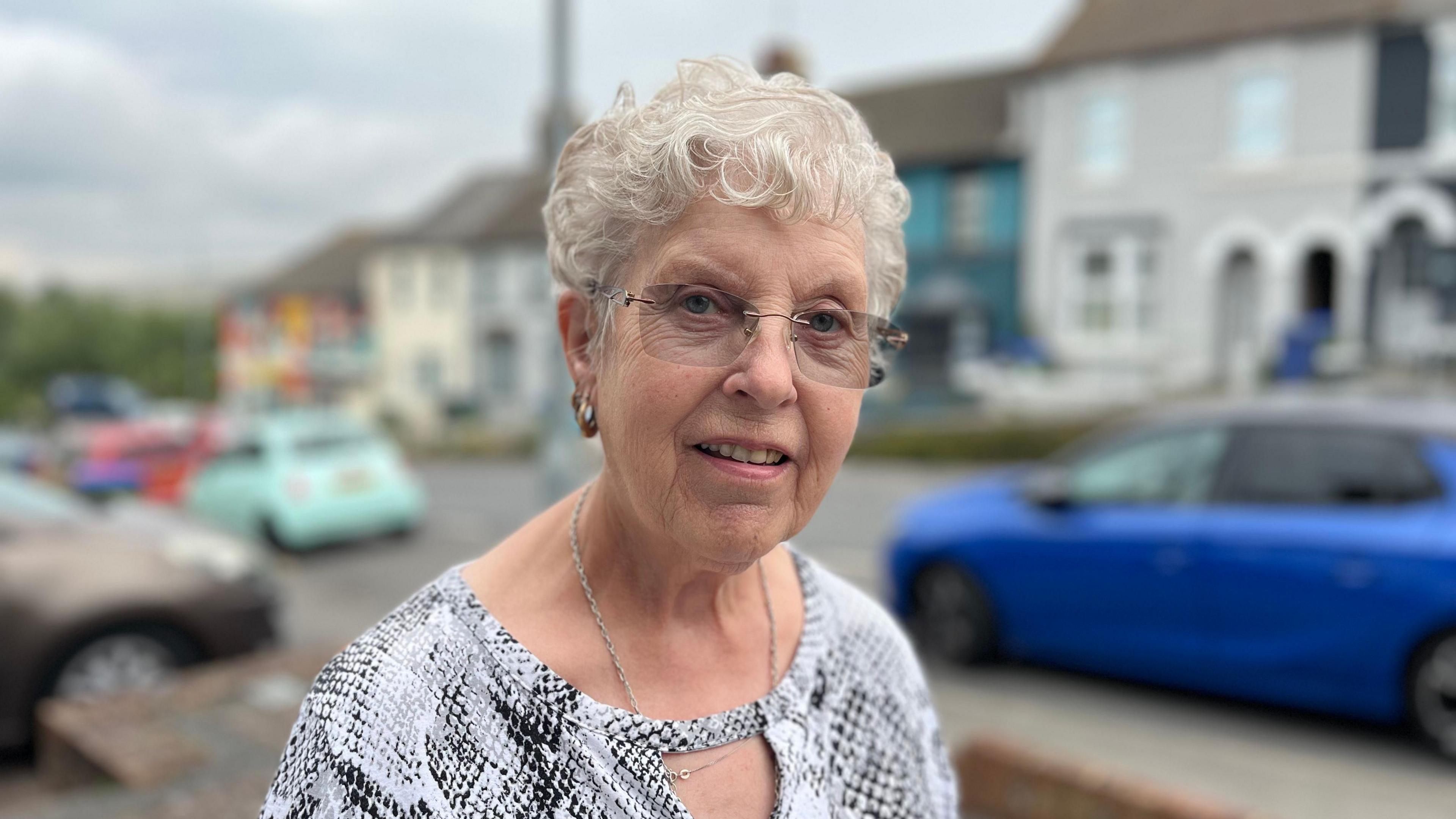 Sylvia Porritt standing next to Lewes Road which has a row of parked cars and terraced houses behind her. She is wearing a silver top and has glasses