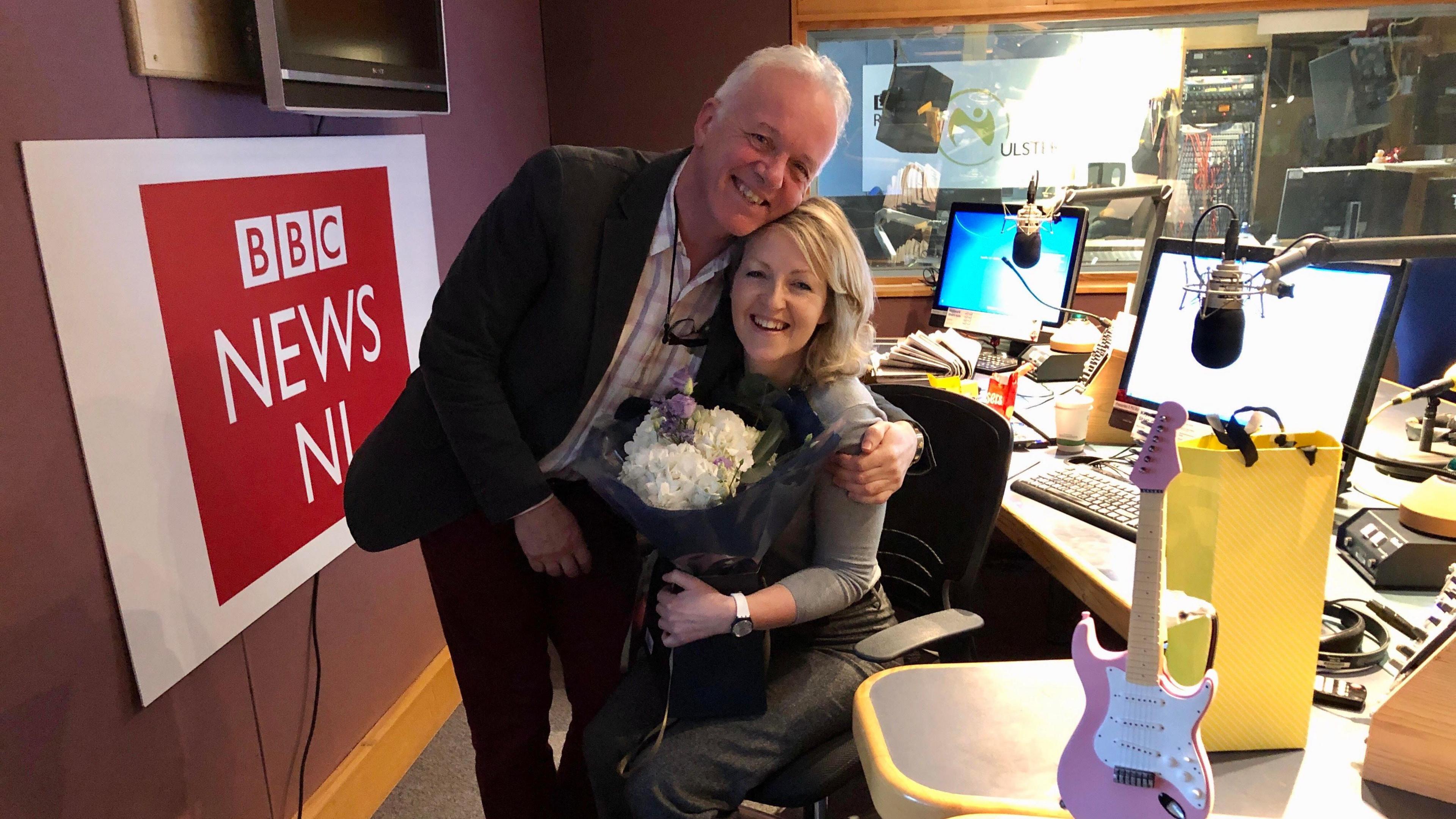 Karen Patterson sitting in a chair in a radio studio holding a bunch of flowers. Noel Thompson is standing next to her, leaning over with his arm round her. To their left a sign with "BBC News NI", to their right a desk with computer screens and microphones