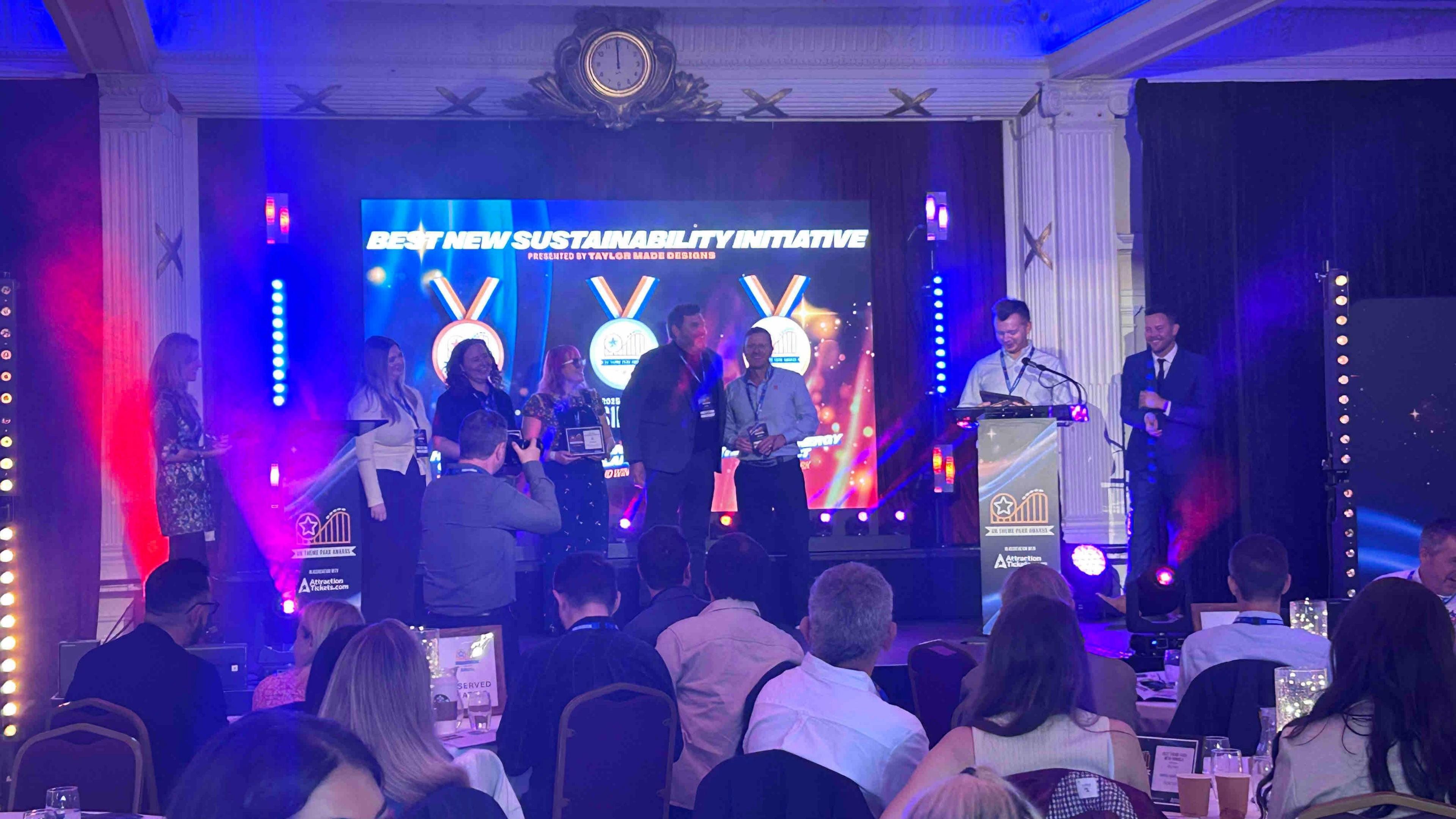 Award ceremony in a decorated hall with a stage and seated audience. People on stage hold awards and speak at podiums.