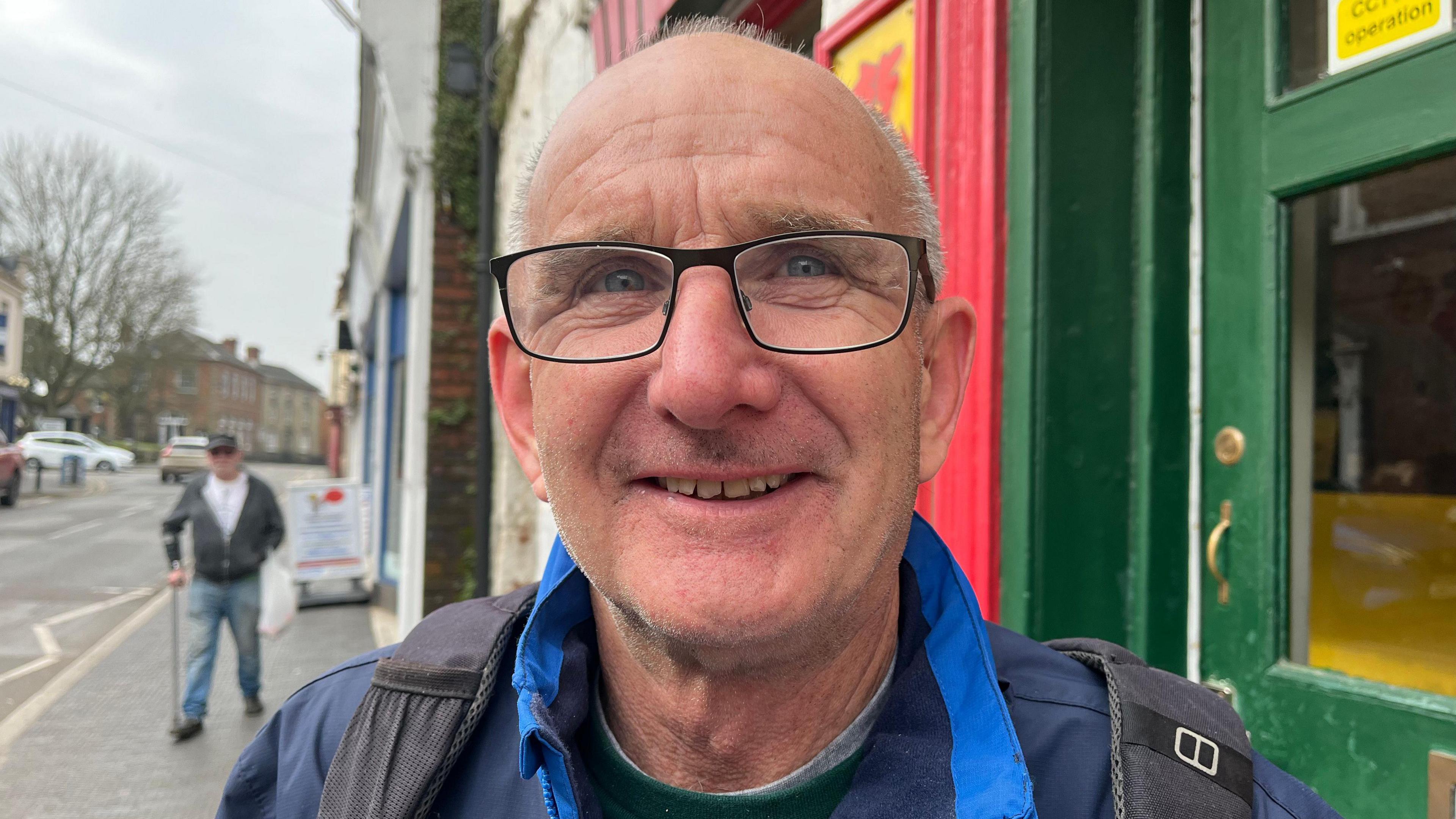 David smiling and looking into the camera; he's wearing a blue waterproof coat with black backpack straps just visible on his shoulders. He's stood on the pavement with a green painted shop window just visible over his left shoulder. Over his right shoulder is a road and more pavement. The sky is grey and overcast.