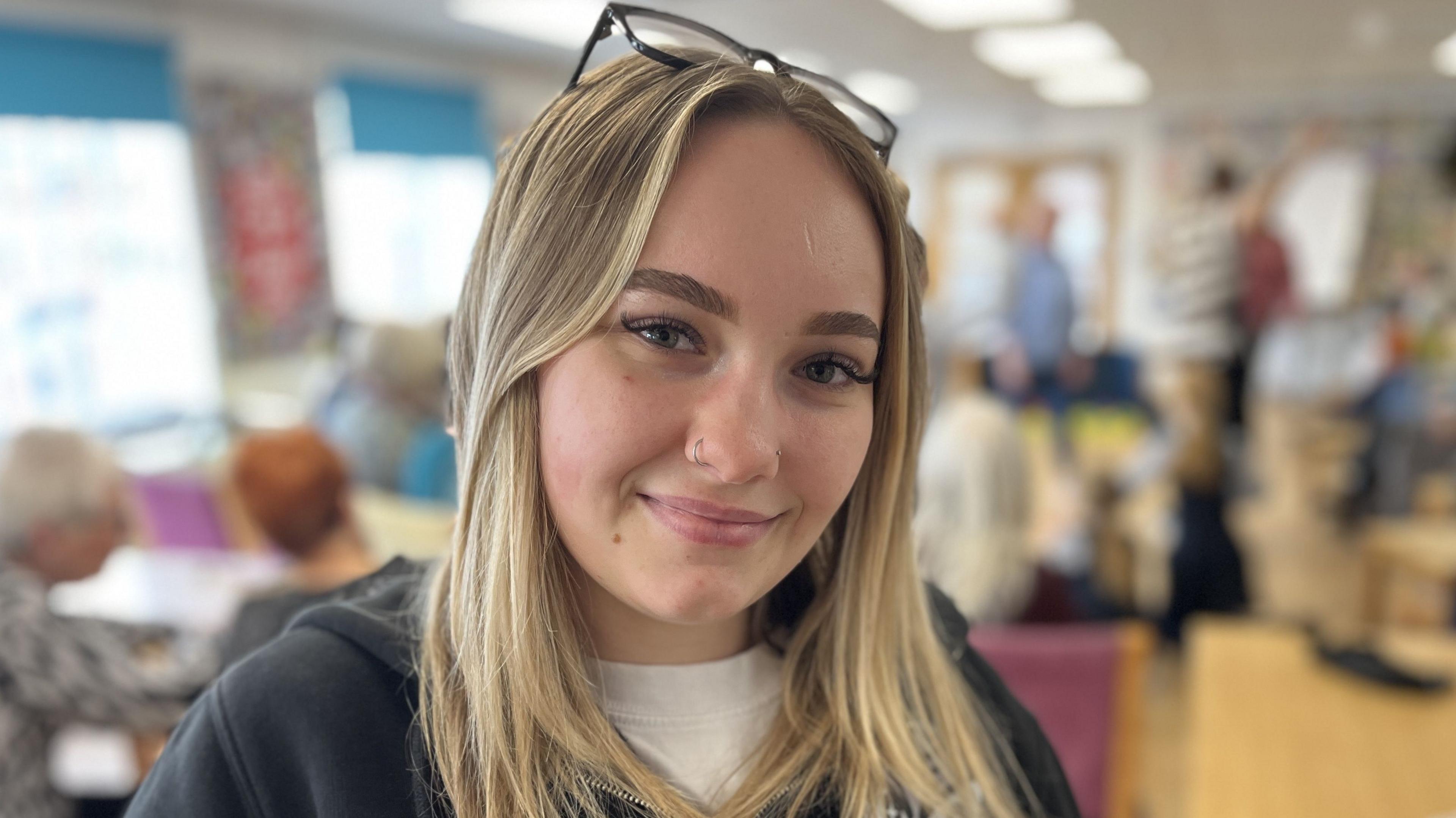 Alex Holton, smiling, with shoulder-length blonde hair, a nose ring and glasses on her head