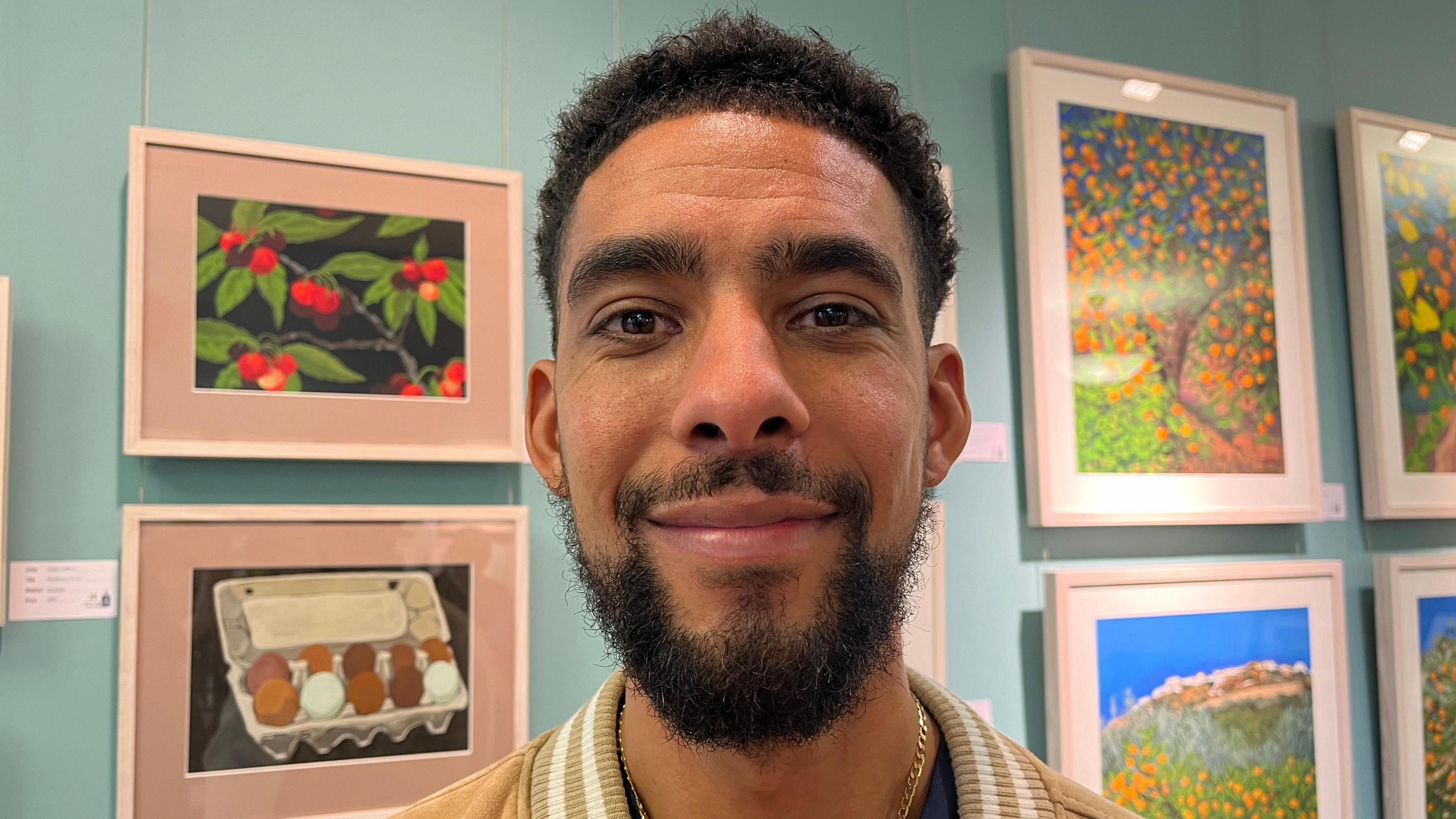 Danny Lloyd-Jones has black hair and a black beard and moustache. He is standing in front of a light blue wall that has photo frames with paintings of food in them