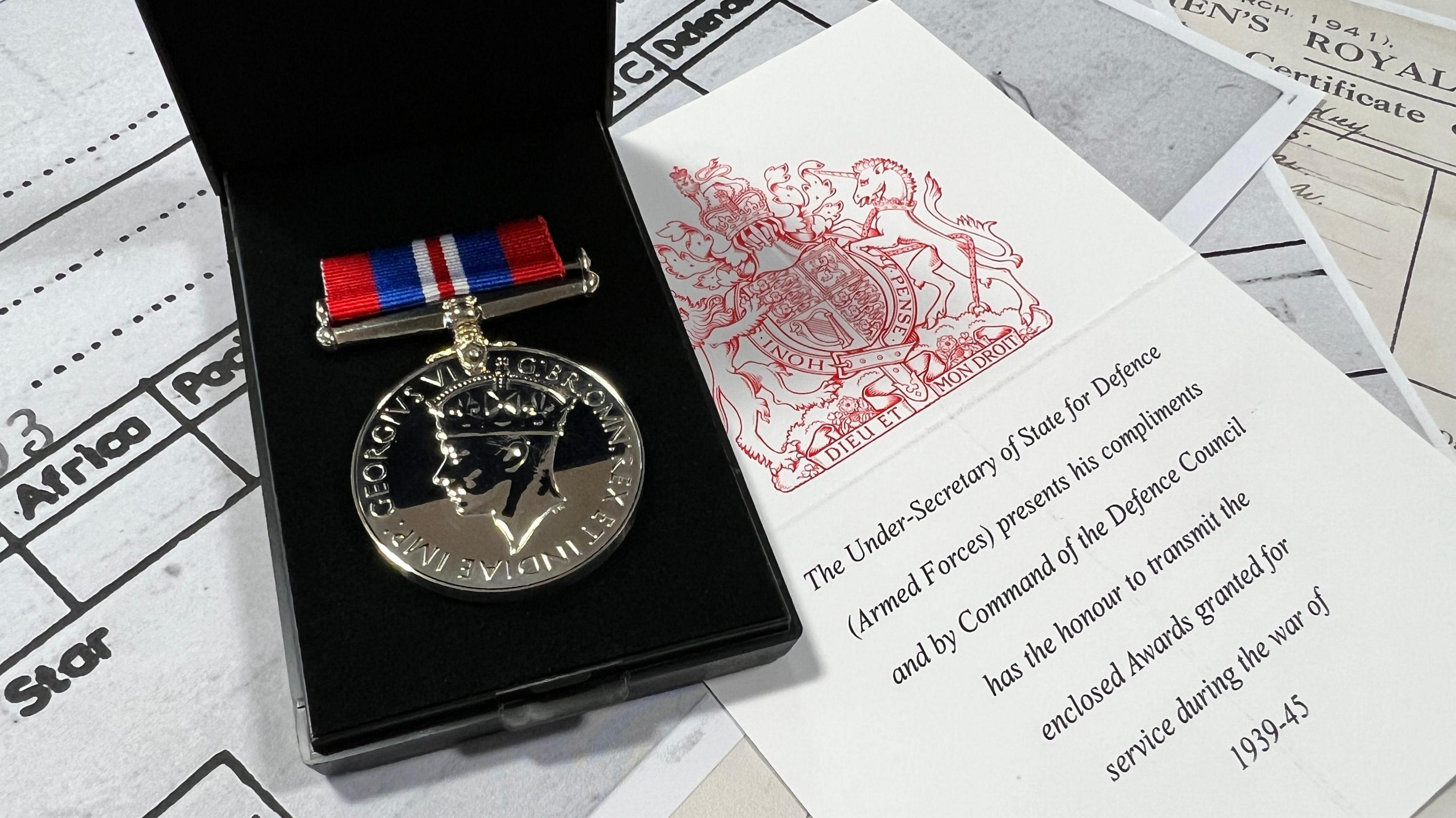 A medal featuring the profile of King George VI, displayed in a black case with a red, white, and blue ribbon, alongside a certificate bearing an embossed red seal and text recognising service during the Second World War