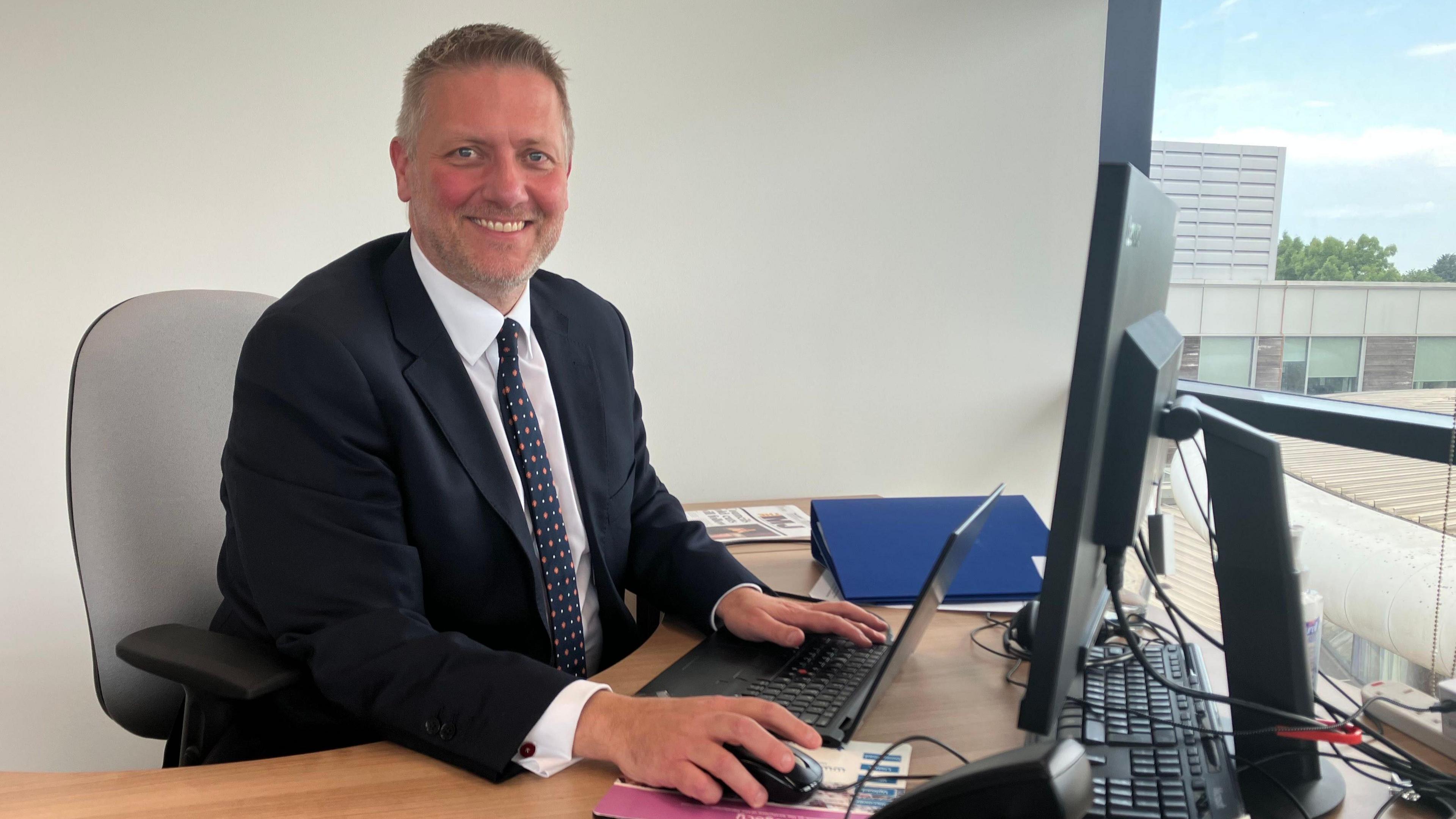 A picture of new Conservative Buckinghamshire council leader Steven Broadbent. He is sat down in front of his laptop and desk, and is wearing a black suit jacket with a white shirt and black tie with red spots.