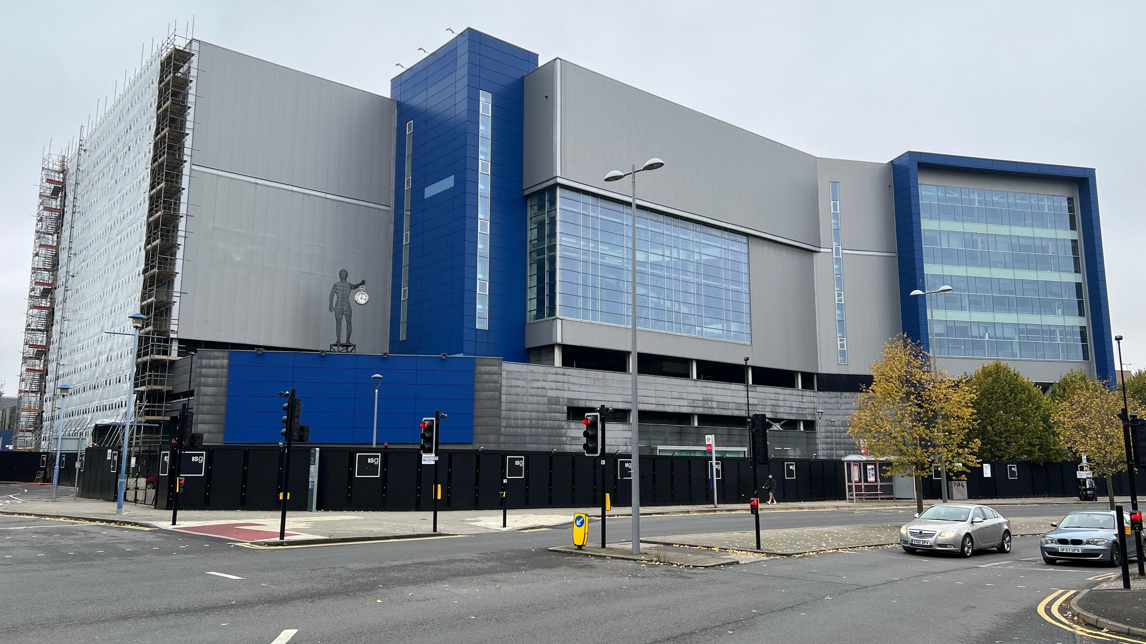 The former Ikea building is pictured, surrounded by scaffolding and construction hoardings. A statue holding a watch is visible in the foreground. The pannelling on the site is blue and grey.