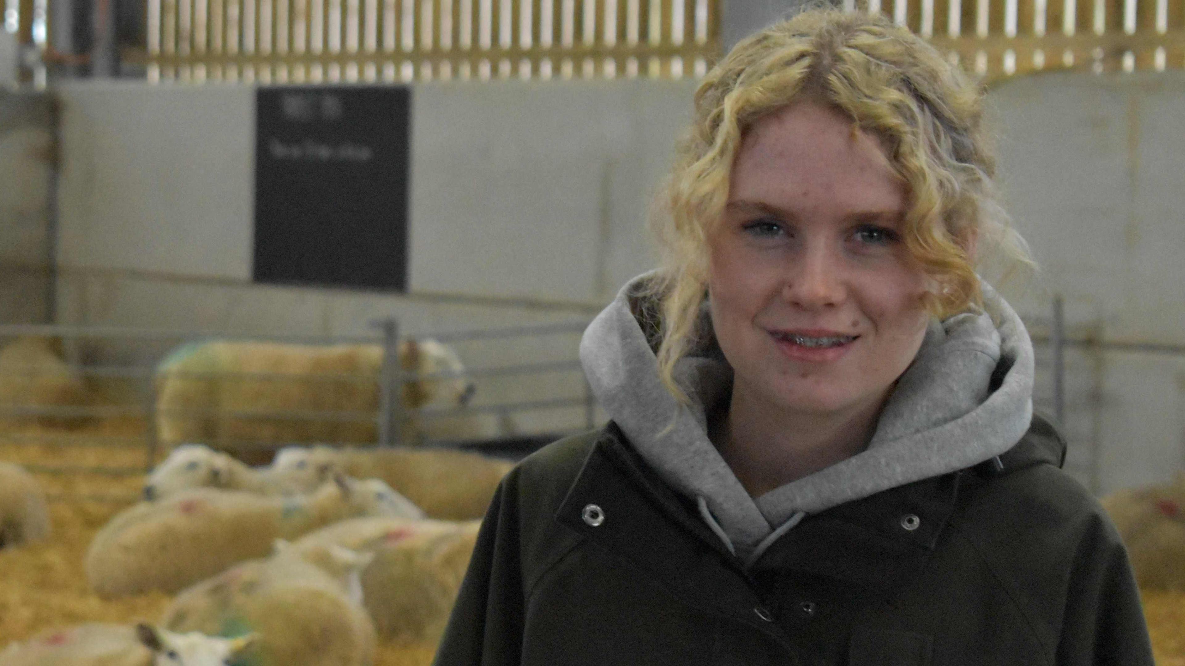 Ellie has curly blonde hair tied back. She has a grey hoodie underneath her black overcoat. Behind her in the barn are sheep sitting on straw