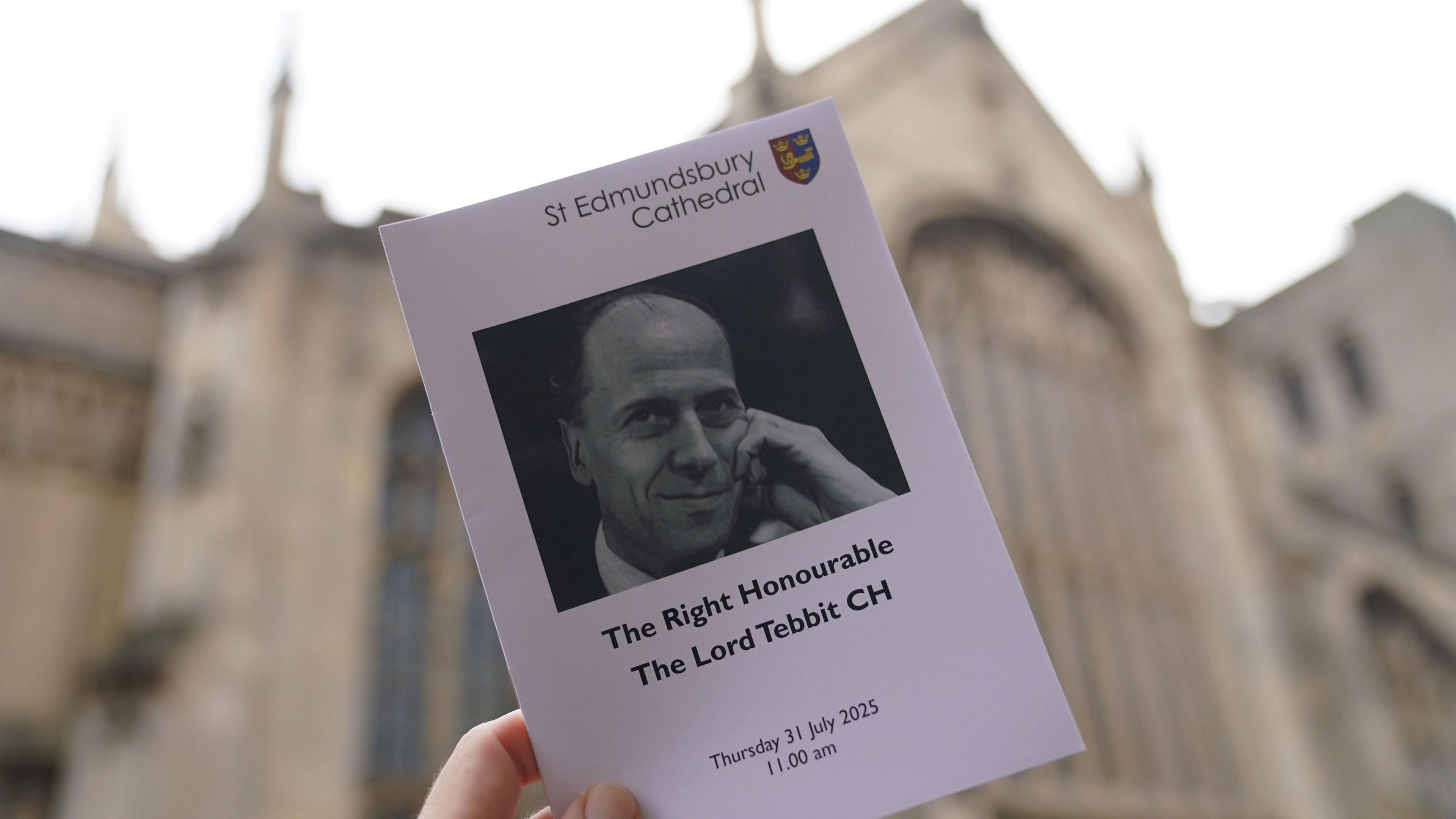 An order of service is held up in front of St Edmundsbury Cathedral, it has a black and white image of Lord Tebbit and details of the date and timings of the funeral service.