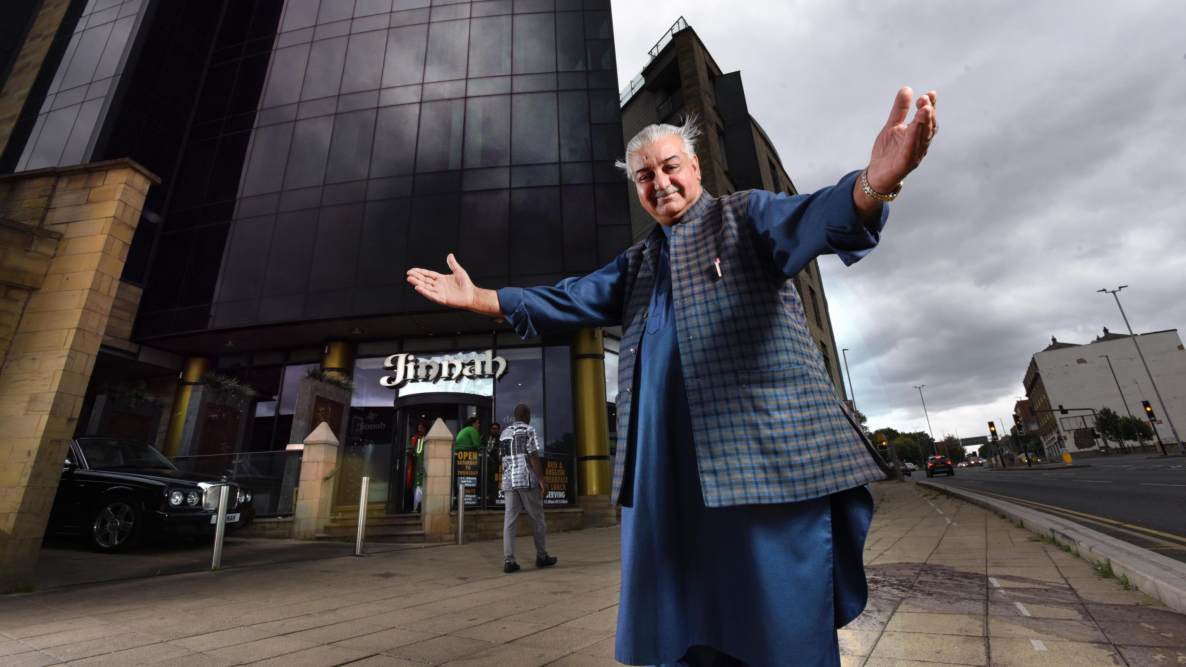 A man stands with arms outstretched, in a welcome pose, outside a restaurant. An urban streetscape is in the background. He wears traditional South Asian dress.