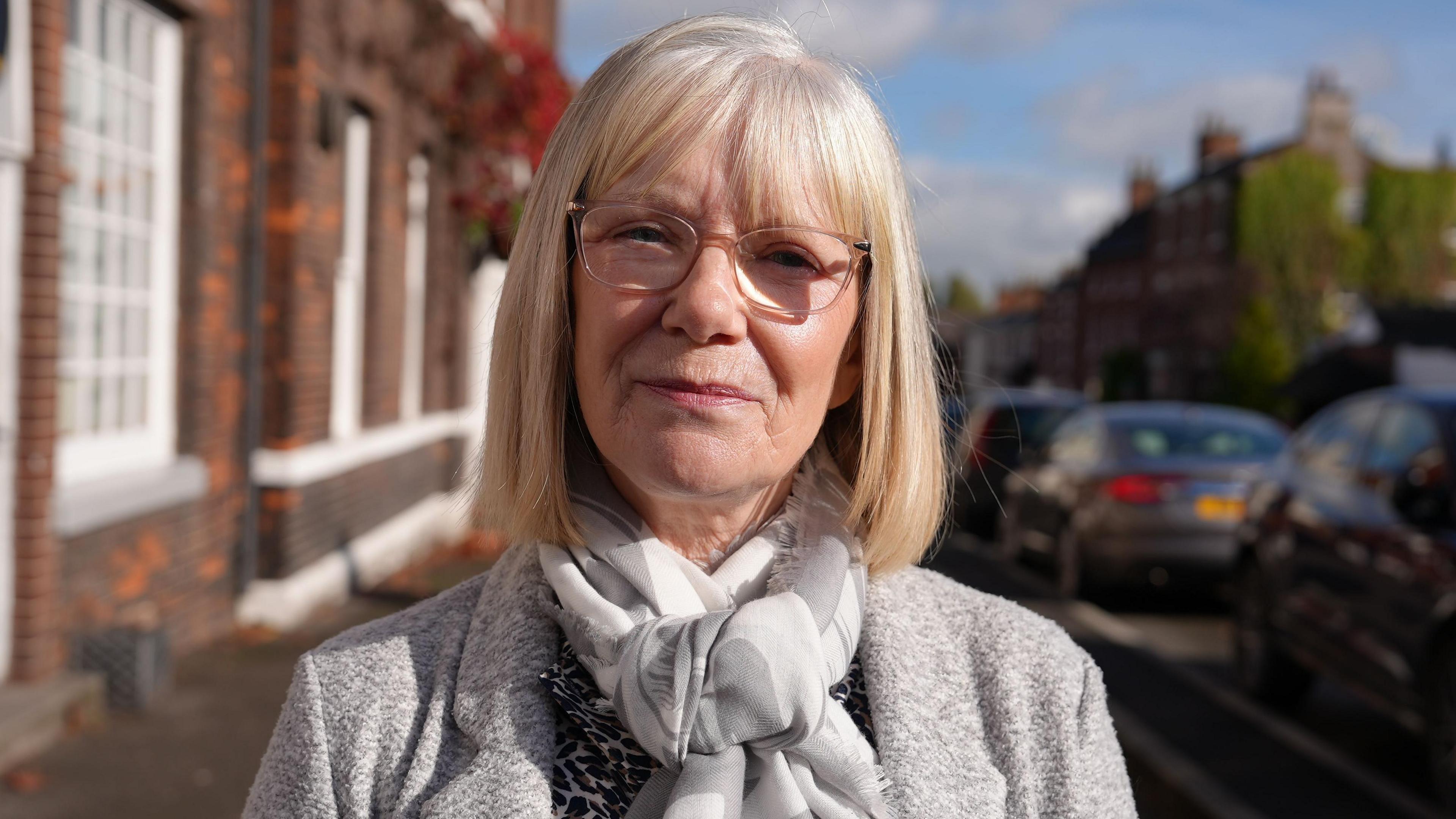 Head and shoulders of a woman with long blonde hair, glasses, a white and grey scarf and a grey coat.